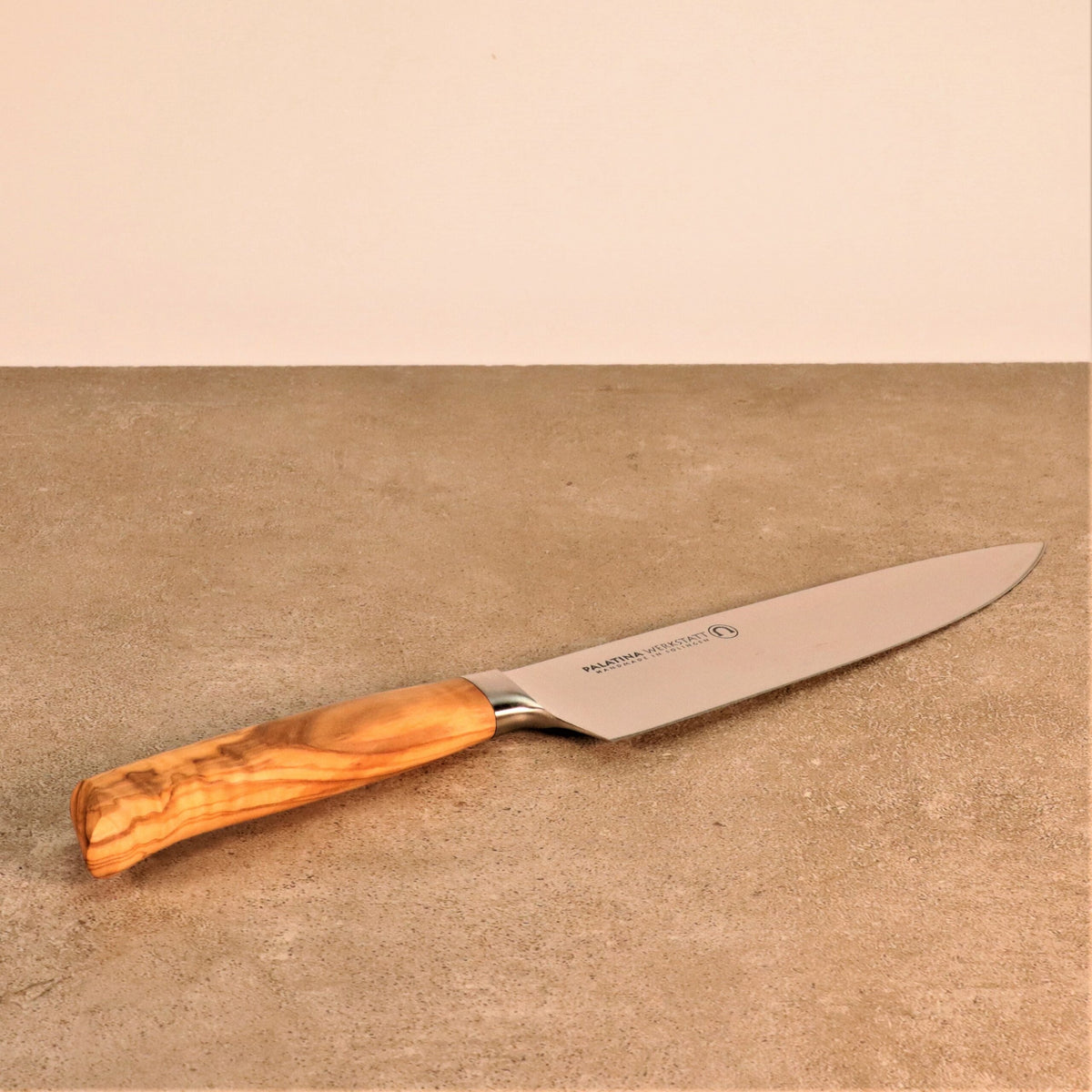 Ein scharfes Küchenmesser mit einem Griff aus alten Fassdauben und Olivenholzgriff liegt auf einer beigen Arbeitsplatte vor einer hellen Wand. Die Klinge besteht aus rostfreiem Stahl und der Griff weist eine natürliche Holzmaserung auf.