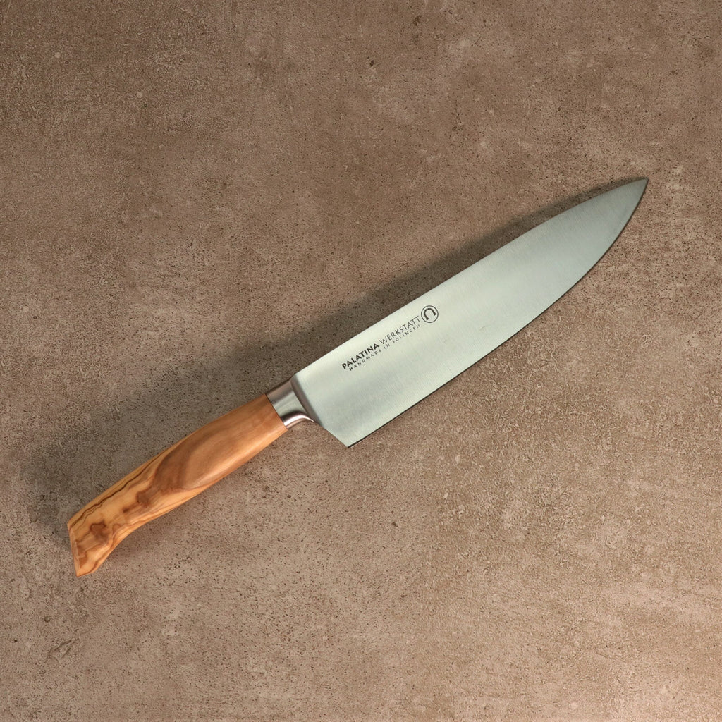 Ein Kochmesser der Palatina Werkstatt Messerserie mit einer 21 cm langen Klinge aus rostfreiem Stahl und einem polierten Olivenholzgriff liegt auf einer strukturierten beigen Oberfläche. Die Klinge weist in der Nähe des Griffs eine Gravur auf.