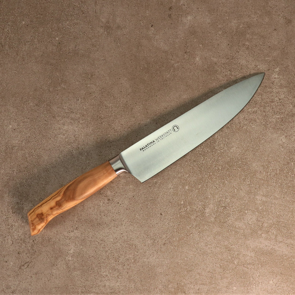 Ein Kochmesser der Palatina Werkstatt Messerserie mit einer 21 cm langen Klinge aus rostfreiem Stahl und einem polierten Olivenholzgriff liegt auf einer strukturierten beigen Oberfläche. Die Klinge weist in der Nähe des Griffs eine Gravur auf.