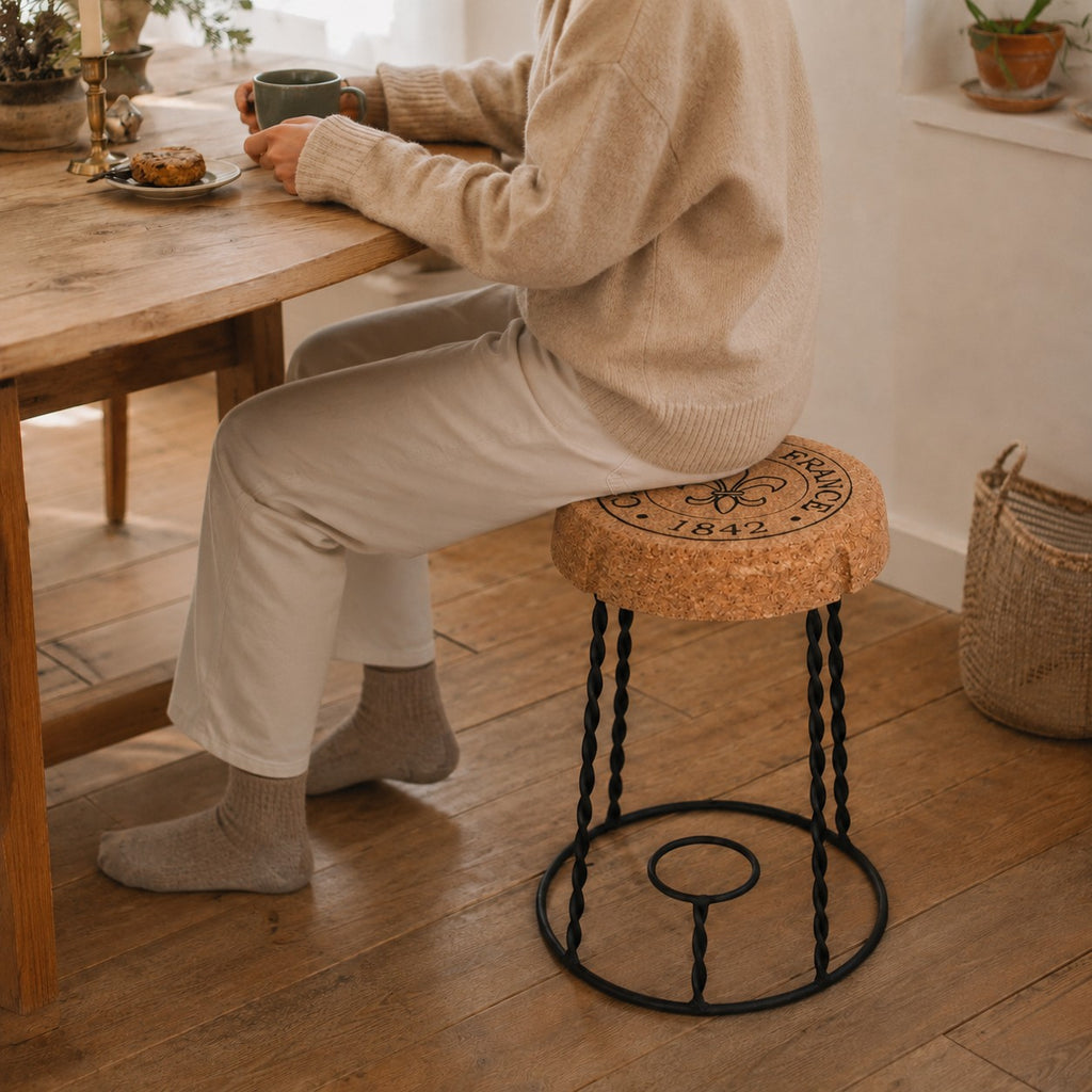 Eine Person in einem beigen Pullover, einer hellen Hose und grauen Socken sitzt auf dem hochwertigen Beistelltisch aus Kork "Agraffe" an einem Holztisch und hält eine Tasse und einen Teller in einem Raum mit Holzboden und gemütlicher, natürlicher Einrichtung.