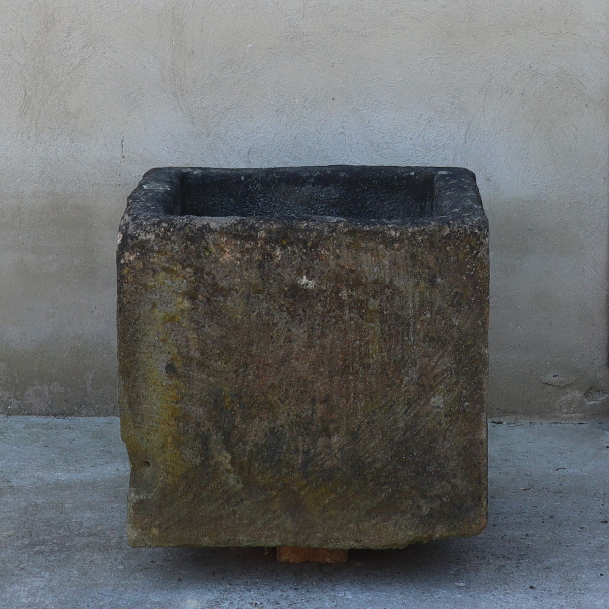 Ein verwitterter antiker Sandsteinkübel (60 x 57 x 53) steht auf einer grauen Betonfläche vor einer strukturierten beigen Wand. Der Blumentopf weist Alterserscheinungen auf und hat eine raue, unebene Oberfläche. Unten befindet sich eine kleine Lücke, die auf einen möglichen Holzblock hindeutet, der ihn stützt.