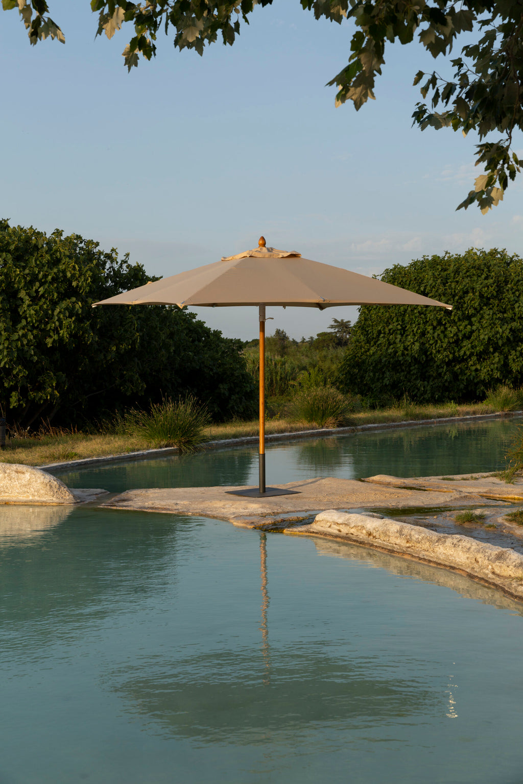 Ein ruhiger Außenpool, umgeben von üppigem Grün. In der Mitte spendet ein großer, beigefarbener Runder Sonnenschirm Lipari (350 - 400 cm) Schatten. Im Wasser des Pools bilden sich sanfte Wellen, die den Sonnenschirm und die umstehenden Bäume reflektieren und eine friedliche und einladende Atmosphäre schaffen, die dem Küstencharme von Lipari ähnelt.