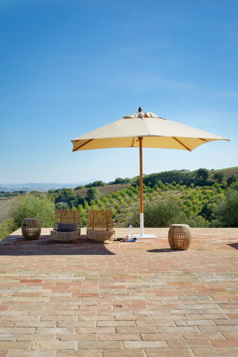 Ein ausladender runder Sonnenschirm Lipari 350 - 400 cm spendet zwei hölzernen Liegestühlen auf einer Terrasse mit Terrakottafliesen Schatten. Neben den Stühlen stehen zwei runde Beistelltische. Die Terrasse mit dem bemerkenswerten Sonnenschirm Lipari bietet einen atemberaubenden Blick auf grüne, sanfte Hügel unter einem klaren blauen Himmel.