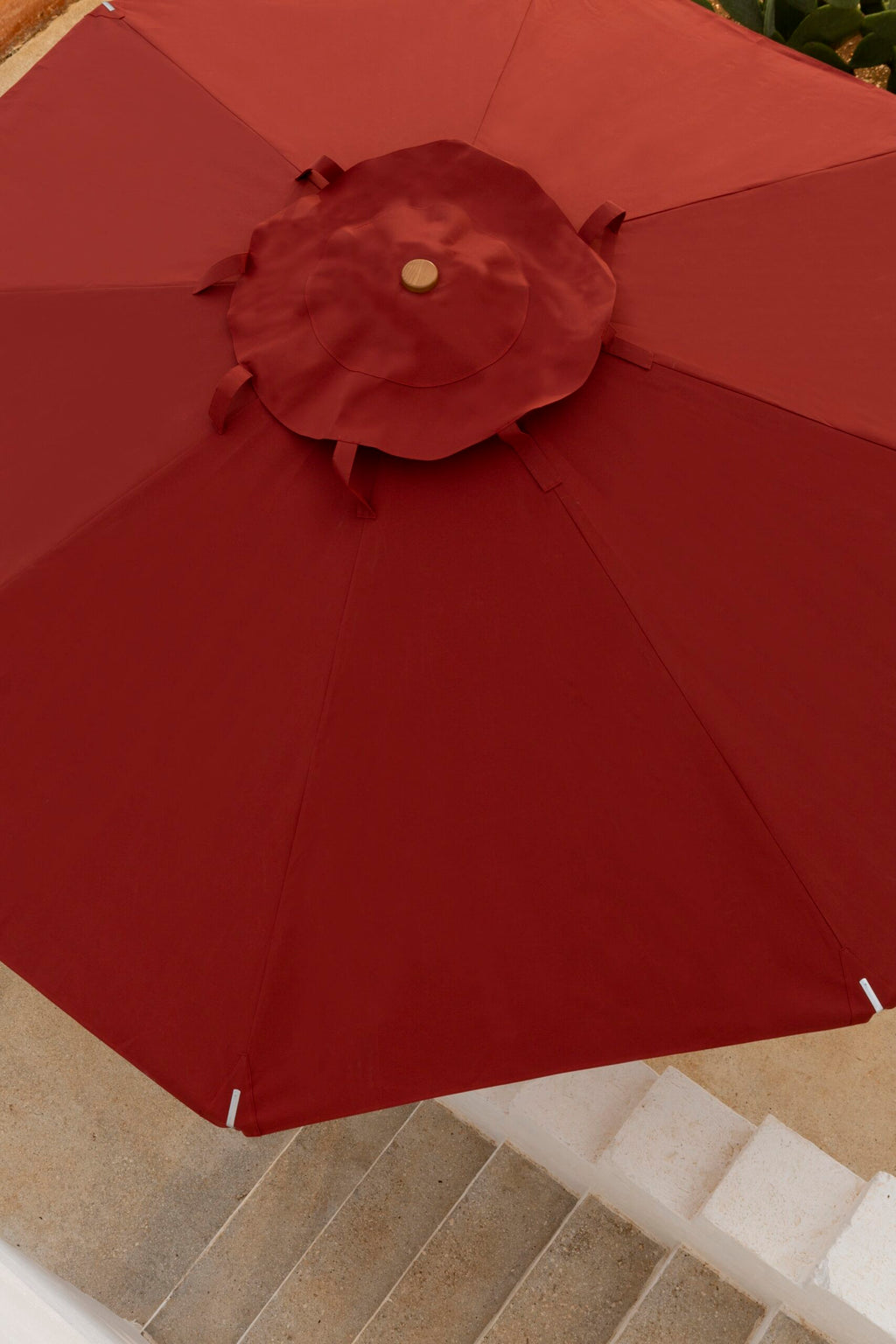 Ein großer runder Sonnenschirm Lipari, 350 - 400 cm groß, mit einem dekorativen zentralen Knotenpunkt ist geöffnet und spendet Schatten über einer weißen Steintreppe. Die leuchtend rote Farbe des Schirms kontrastiert mit den hellen Stufen und dem umgebenden Grün.