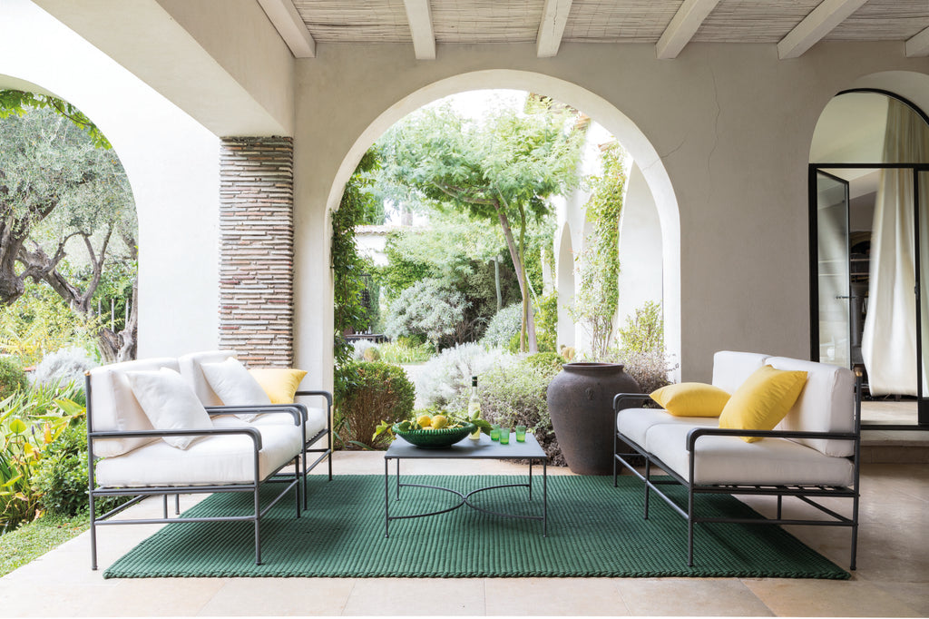 Außenterrasse mit einem graphitgrauen Sessel Toscana aus Eisen mit weißen Kissen, gelben und weißen Zierkissen, einem Couchtisch mit Glasplatte und einem grünen strukturierten Teppich. Die Terrasse verfügt über gewölbte Öffnungen, die einen Blick auf üppiges Grün bieten, das an die Toskana erinnert, ergänzt durch eine Steinsäule und hohe Topfpflanzen in der Nähe.