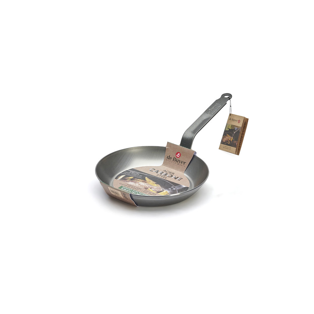 Die De Buyer Lyonnaise Bratpfanne Carbone Plus ist eine Metallpfanne mit einem langen Griff, einem Etikett aus Pappe und einem braunen Anhänger. Der Markenname "de Buyer" steht auf dem Etikett im Inneren der Pfanne.