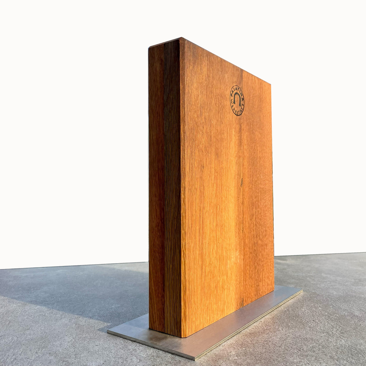 Ein großes Holzbuch, gefertigt aus den Dauben alter Weinfässer und mit glatter Oberfläche, steht aufrecht auf einem Sockel aus Edelstahl. Die Rückseite zeigt einen kreisförmigen Stempel mit einer teilweise sichtbaren Gravur. Das Buch steht vor einem minimalistischen Hintergrund mit einer grauen Oberfläche und einer weißen Wand.