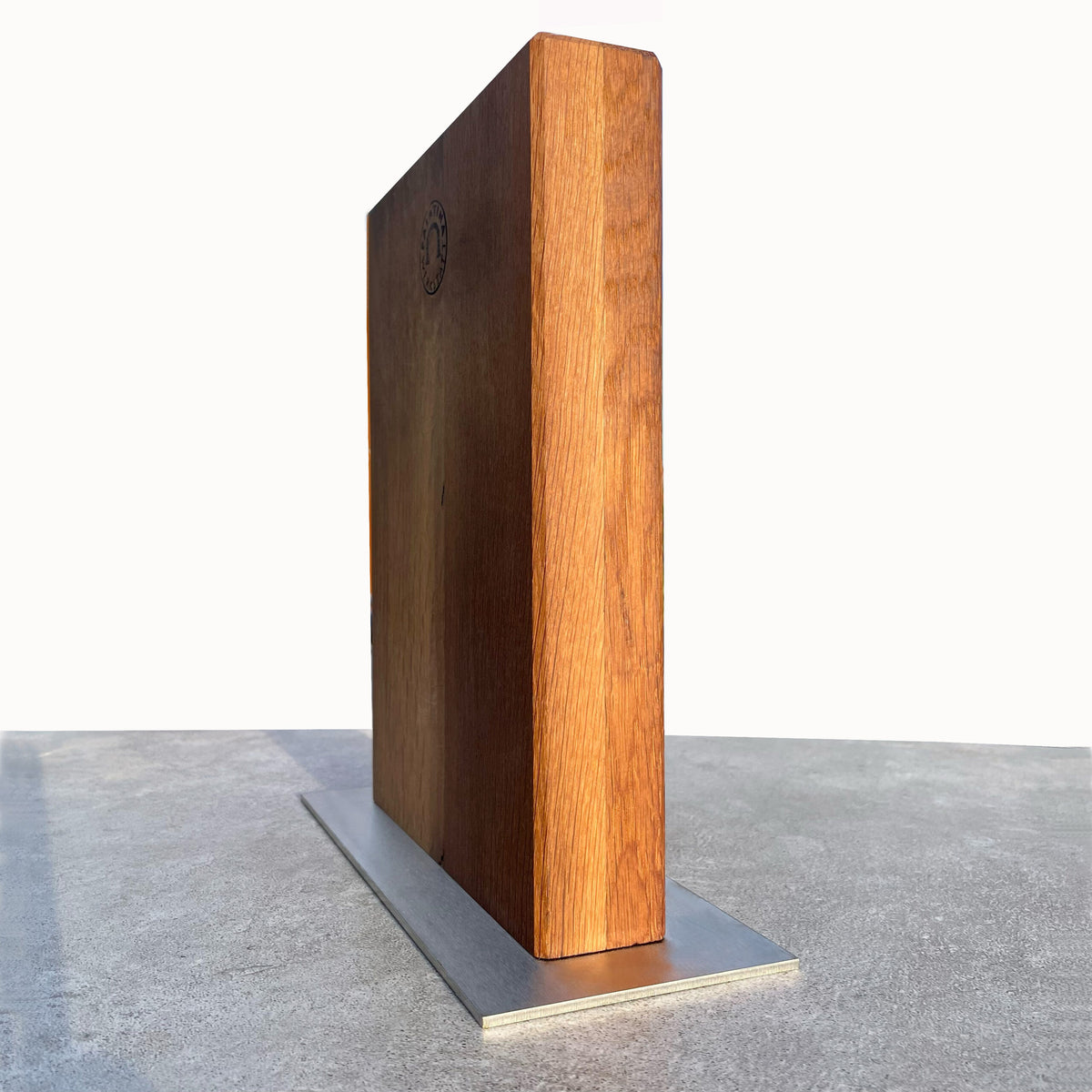 Das Bild zeigt den Palatina Werkstatt Magnet-Messerblock aus Fassdauben von alten Weinfässern, vertikal positioniert auf einem Edelstahlsockel. Das Holz hat eine reiche, natürliche Maserung und auf der Oberseite befindet sich ein kleines eingraviertes Emblem. Der metallische Sockel wirkt flach und robust und bietet dem Messerblock stabilen Halt.