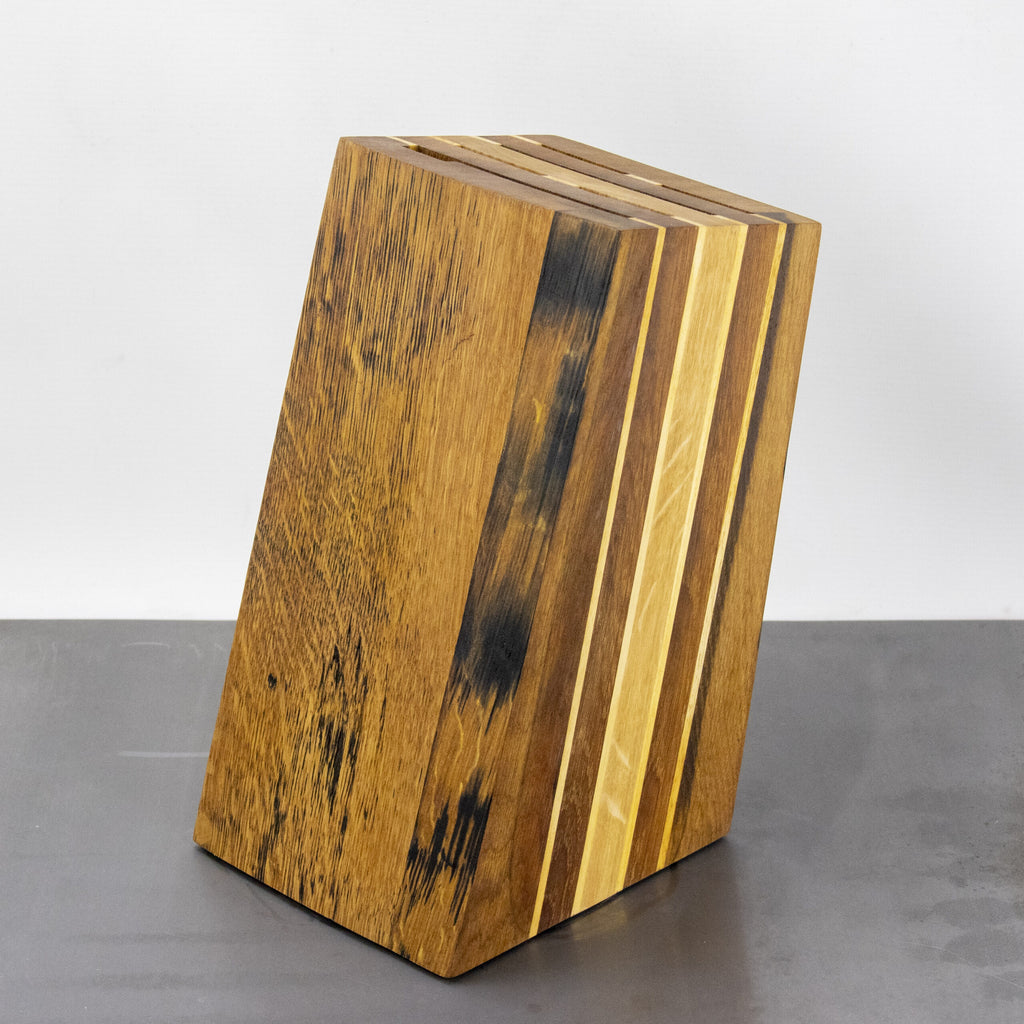 Der schräge Messerblock aus den Fassdauben von alten Eichen-Weinfässern von Palatina Werkstatt weist ein leicht schräges Design auf und zeigt ein natürliches Holzmaserungsmuster mit abwechselnd hellen und dunklen Holzstreifen. Er hat oben mehrere Schlitze zum Halten von Messern, die sich vor einem schlichten, hellen Hintergrund abheben.