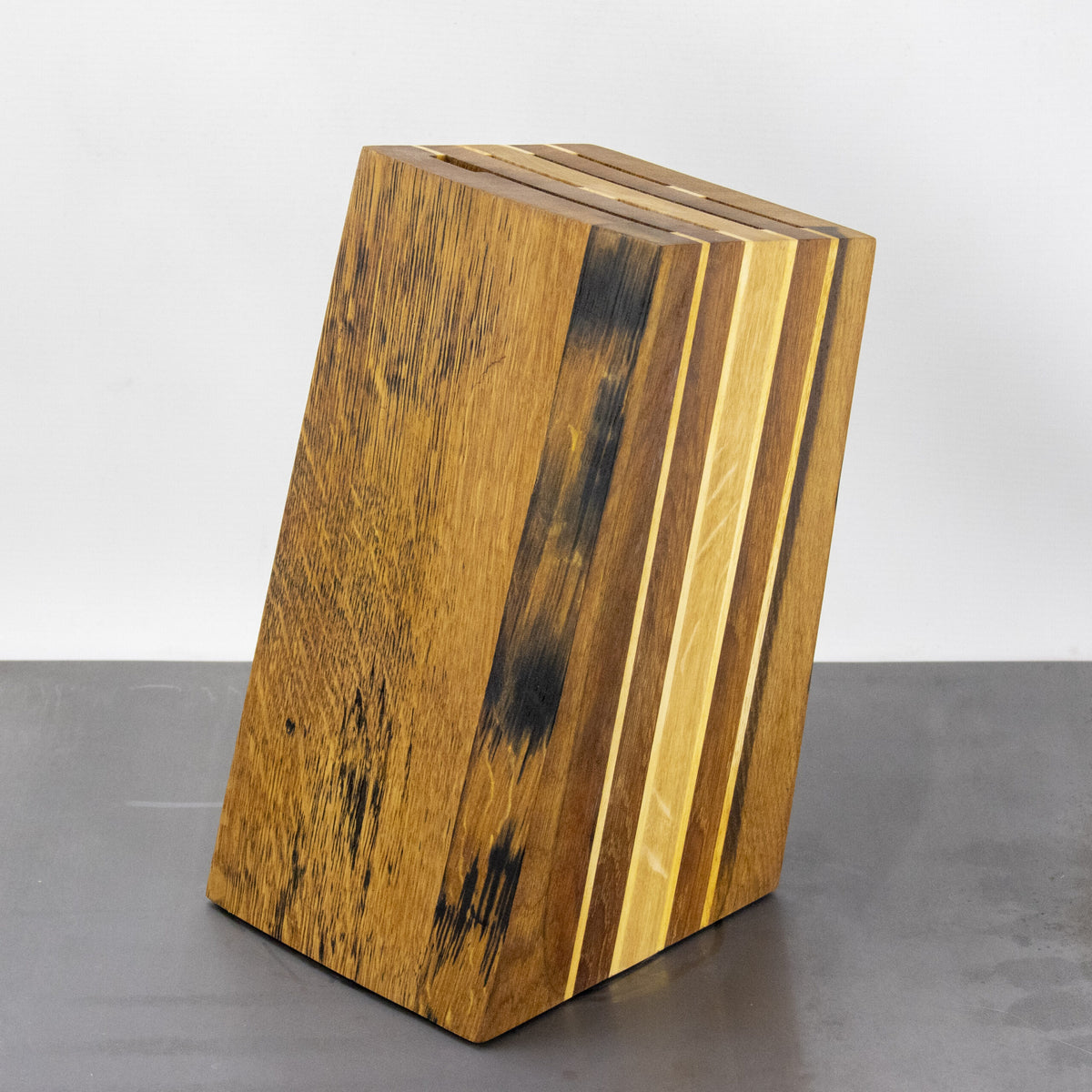 Der schräge Messerblock aus den Fassdauben von alten Eichen-Weinfässern von Palatina Werkstatt weist ein leicht schräges Design auf und zeigt ein natürliches Holzmaserungsmuster mit abwechselnd hellen und dunklen Holzstreifen. Er hat oben mehrere Schlitze zum Halten von Messern, die sich vor einem schlichten, hellen Hintergrund abheben.