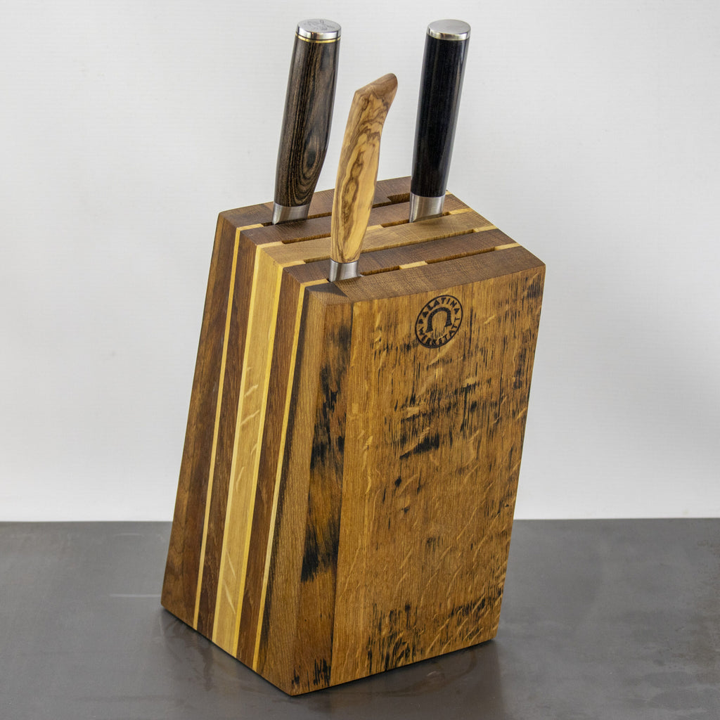 Ein hölzerner Messerblock mit rustikalem Finish, der drei Messer von Palatina Werkstatt enthält. Die Griffe der Messer variieren in Farbe und Muster, wobei einer aus Holz und die anderen dunkler sind und an alte Eichenweinfässer erinnern. Der schräge Messerblock aus den Fassdauben von alten Eichenweinfässern weist auf der Vorderseite ein Markenemblem auf einem schlichten Hintergrund auf.