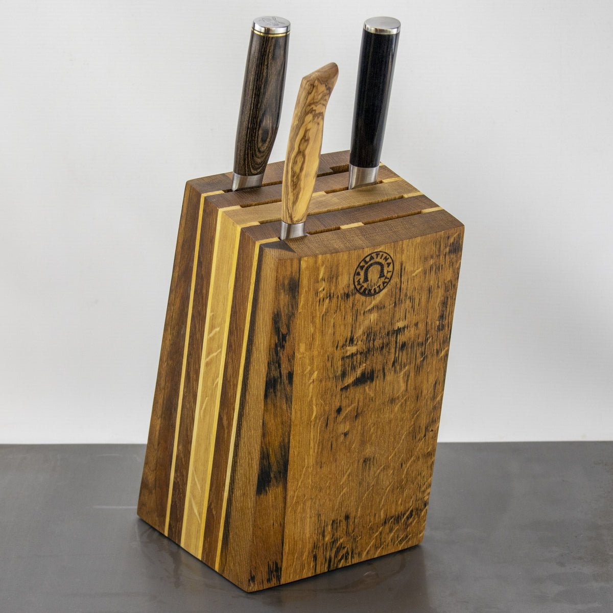 Ein hölzerner Messerblock mit rustikalem Finish, der drei Messer von Palatina Werkstatt enthält. Die Griffe der Messer variieren in Farbe und Muster, wobei einer aus Holz und die anderen dunkler sind und an alte Eichenweinfässer erinnern. Der schräge Messerblock aus den Fassdauben von alten Eichenweinfässern weist auf der Vorderseite ein Markenemblem auf einem schlichten Hintergrund auf.