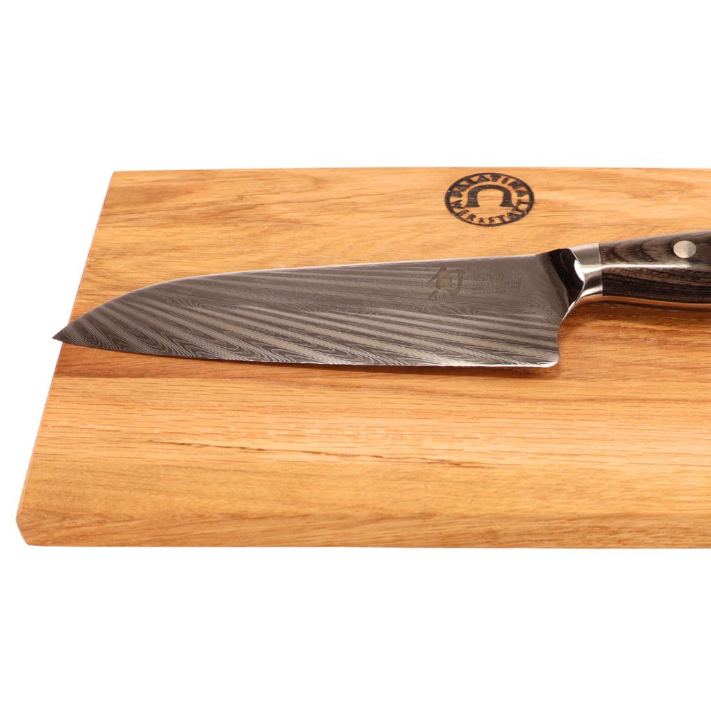 Das KAI Shun Nagare NDC-0702 Santoku-Messer (18 cm, handgeschmiedet) mit gemusterter Klinge und dunklem Griff liegt auf dem 30x20 cm großen Eiche-Schneidebrett mit rundem Logo in der oberen rechten Ecke.