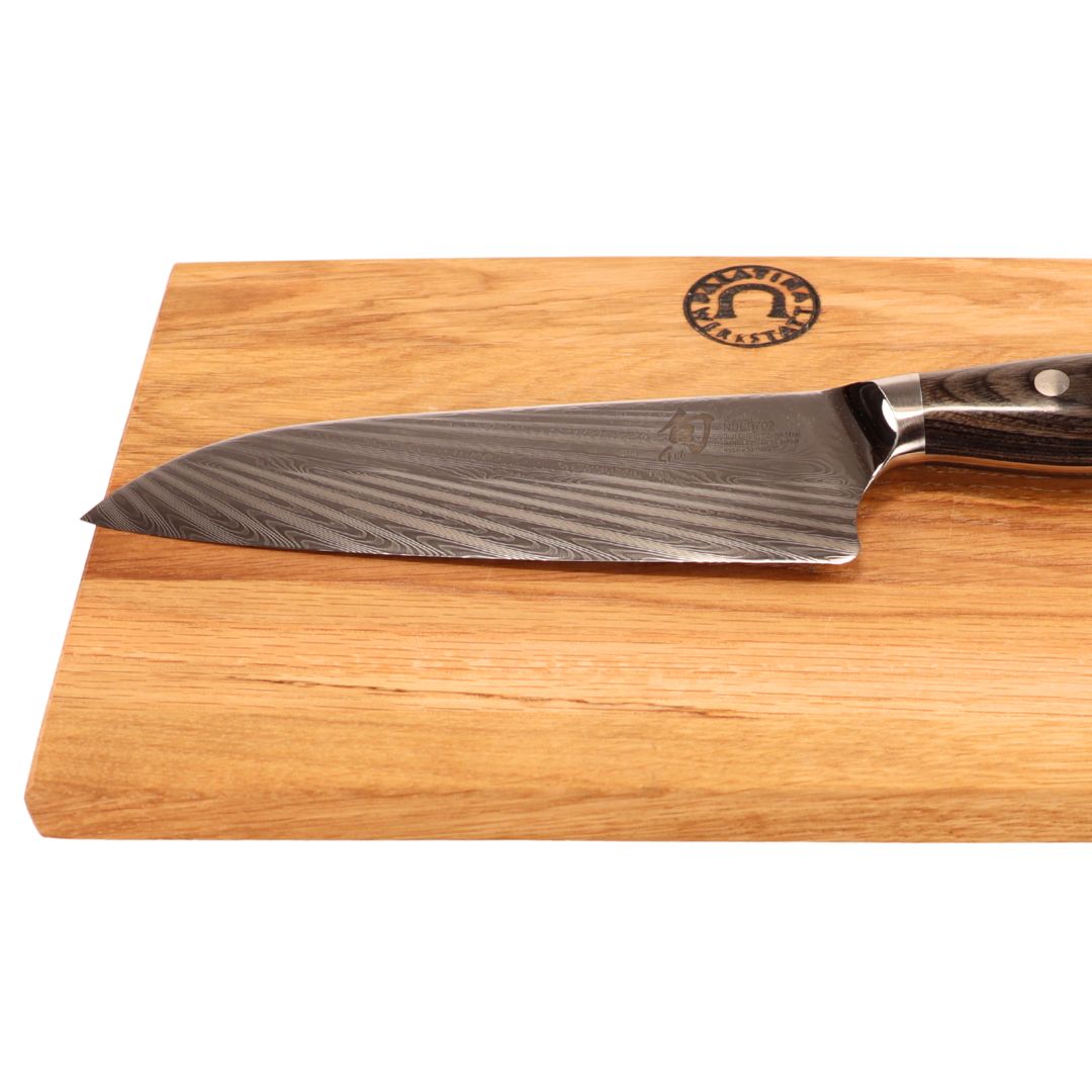 Das KAI Shun Nagare NDC-0702 Santoku-Messer (18 cm, handgeschmiedet) mit gemusterter Klinge und dunklem Griff liegt auf dem 30x20 cm großen Eiche-Schneidebrett mit rundem Logo in der oberen rechten Ecke.