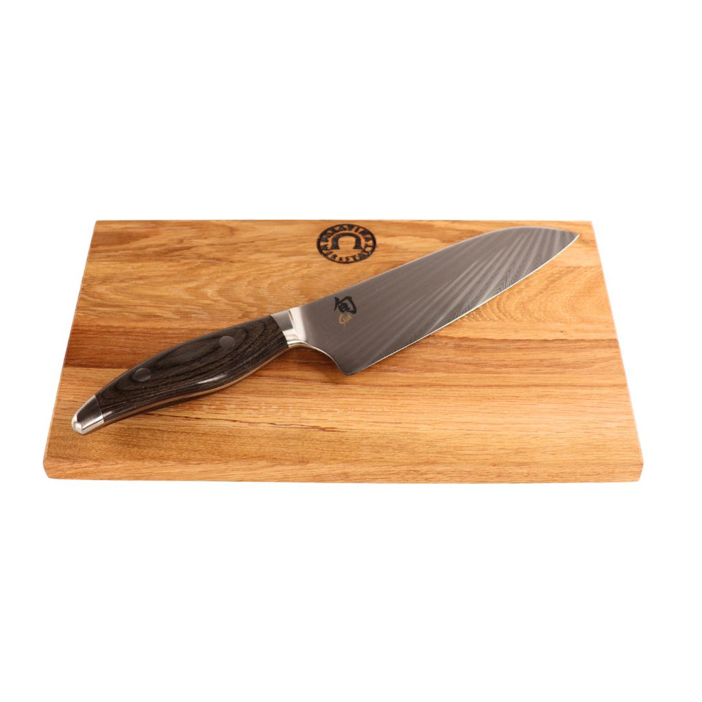 Das KAI Shun Nagare NDC-0702 Santoku-Messer mit 18 cm Klinge und dunklem Griff liegt auf dem 30x20 cm großen Schneidebrett aus Eiche, das oben mittig ein kleines rundes Logo trägt.