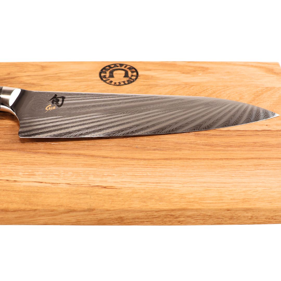 Das Kai Shun Nagare NDC-0706 Kochmesser (72 Lagen Damast, 20 cm) mit gemusterter Klinge liegt auf einem 30x20 cm basaltgrauen Schneidebrett aus Eiche mit rundem Logo.