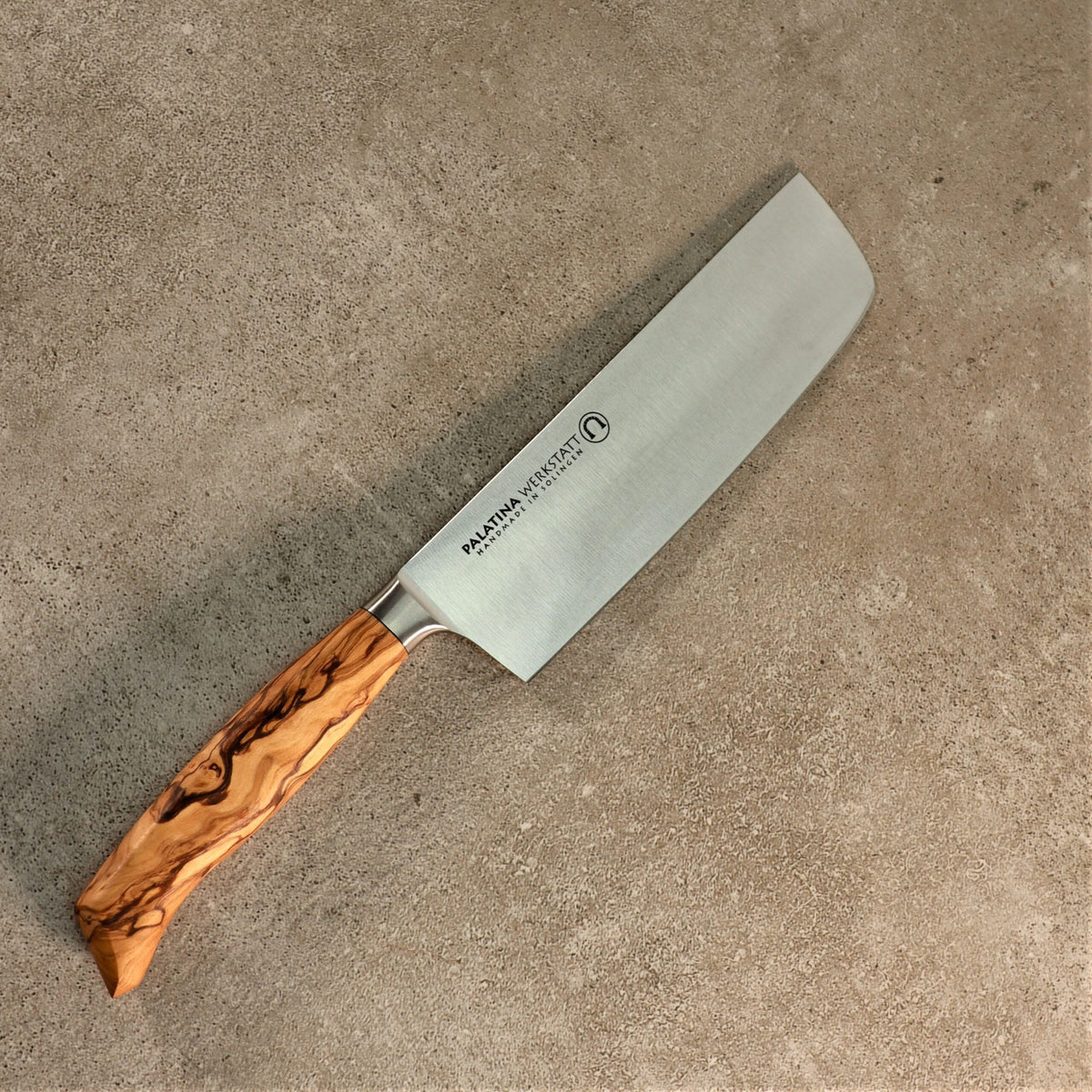 Ein Messer aus der exklusiven, bestückten Messerblock-Kollektion aus alten Fassdauben + 6 Messer liegt auf einer beigefarbenen, strukturierten Oberfläche. Das Küchenmesser hat eine breite, rechteckige Klinge mit dem Markennamen „Makami Knives“ und seinem Logo darauf. Der elegante Griff aus Olivenholz weist eine ausgeprägte, natürliche Holzmaserung auf.