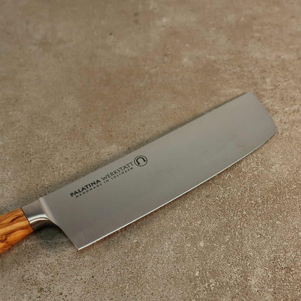 Ein Nakirimesser aus der Palatina Werkstatt Messerserie, mit einer 16 cm breiten silbernen Klinge und einem Olivenholzgriff, liegt auf einer strukturierten grauen Oberfläche. Die Klinge ist mit der Aufschrift „Palatina Werkstatt“ versehen und mit einem kreisrunden Logo verziert.