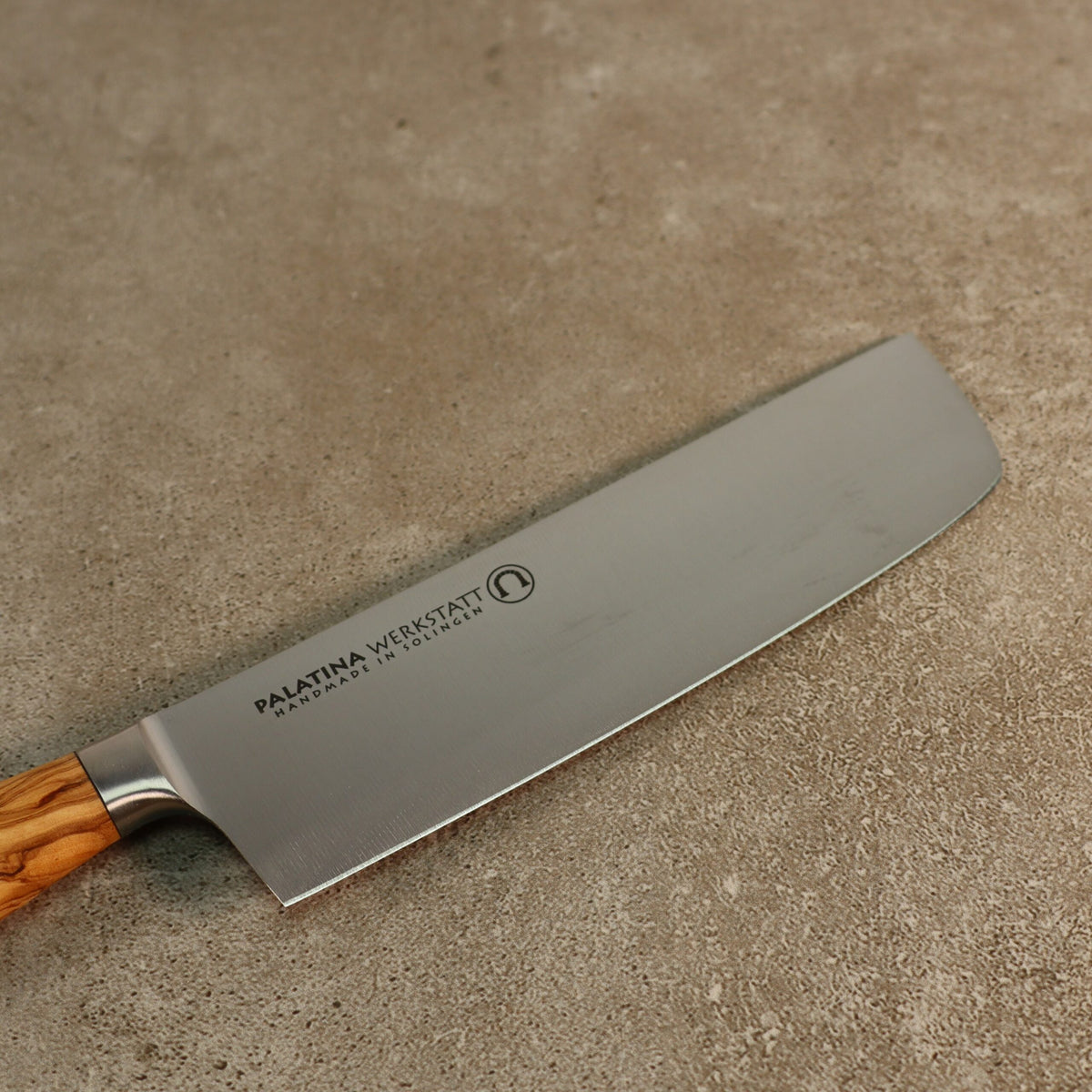 Ein Nakirimesser aus der Palatina Werkstatt Messerserie, mit einer 16 cm breiten silbernen Klinge und einem Olivenholzgriff, liegt auf einer strukturierten grauen Oberfläche. Die Klinge ist mit der Aufschrift „Palatina Werkstatt“ versehen und mit einem kreisrunden Logo verziert.