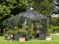 Eine freistehende Orangerie steht bescheiden in einem üppigen, grünen Garten. Im Inneren gedeihen verschiedene Pflanzen und Blumen, während im Außenbereich zusätzliche Topfpflanzen und Gartengeräte aufgestellt sind. Hohe Bäume und kräftiges Grün umgeben die freistehende Orangerie und schaffen einen ruhigen Außenbereich.