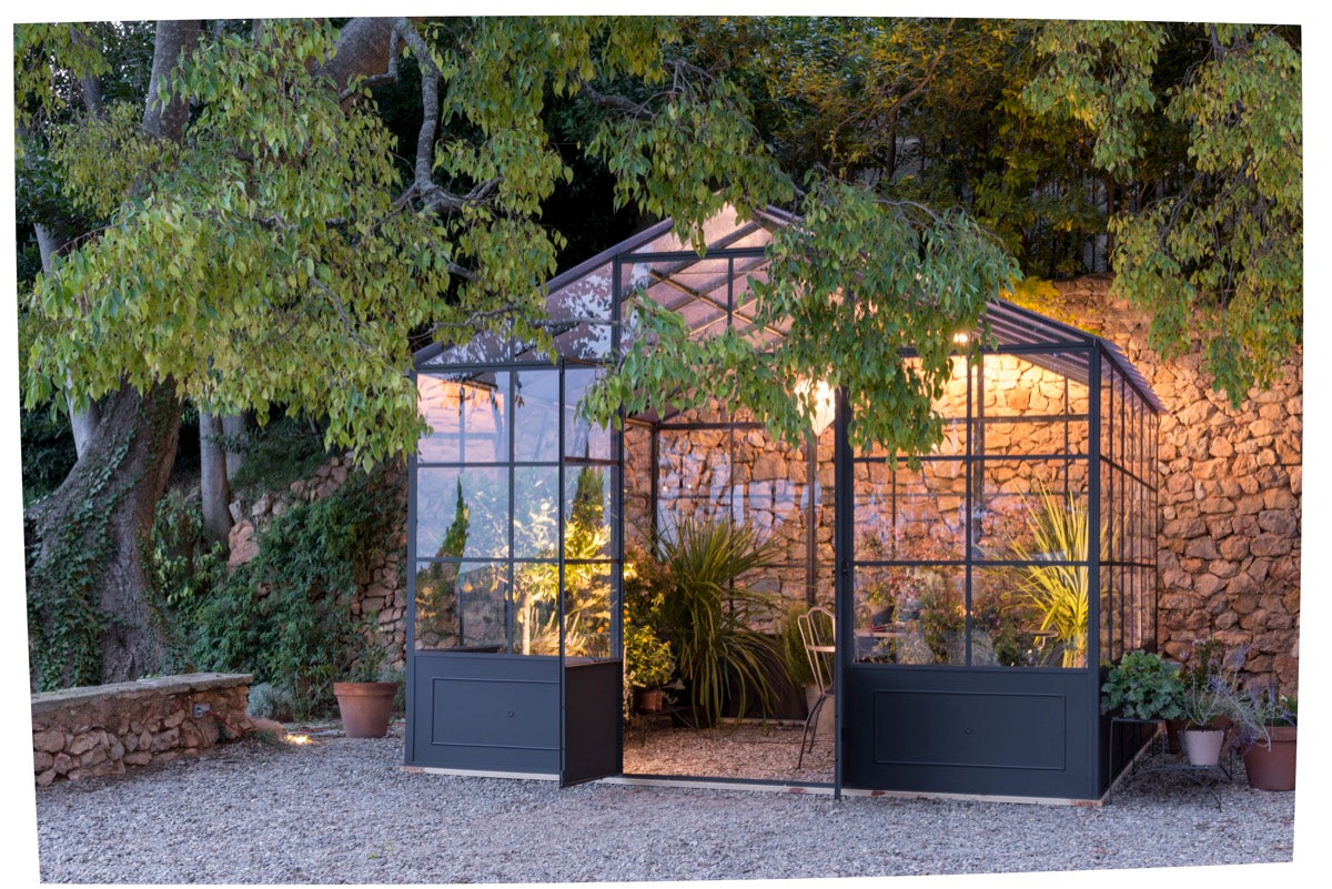 Eine kleine freistehende Orangerie aus Glas ist von üppigen grünen Bäumen und einer Steinmauer umgeben. Der warm beleuchtete Innenraum ist mit verschiedenen Pflanzen gefüllt, und zwei Stühle stehen inmitten des Grüns und schaffen eine gemütliche und einladende Atmosphäre in diesem freistehenden Gewächshaus.