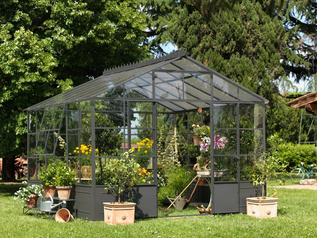 Eine freistehende Orangerie steht bescheiden in einem üppigen, grünen Garten. Im Inneren gedeihen verschiedene Pflanzen und Blumen, während im Außenbereich zusätzliche Topfpflanzen und Gartengeräte aufgestellt sind. Hohe Bäume und kräftiges Grün umgeben die freistehende Orangerie und schaffen einen ruhigen Außenbereich.