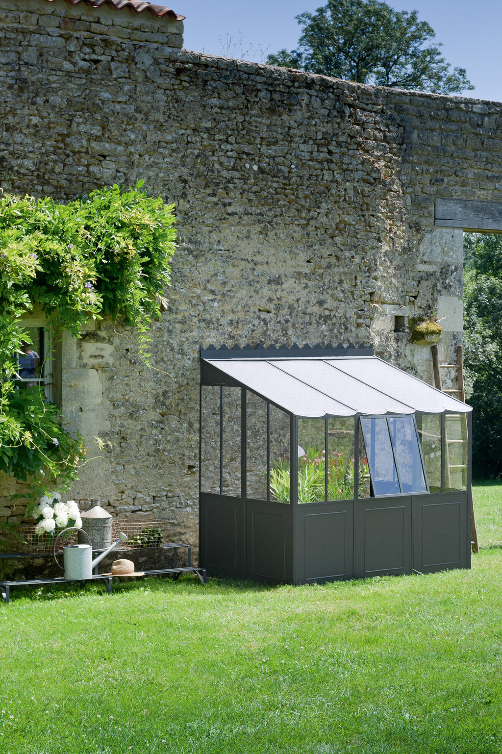 Eine kleine freistehende Orangerie mit Glasscheiben und dunklem Rahmen steht vor einer alten Steinmauer, die teilweise von Grün bedeckt ist. Das Gewächshaus steht auf einem üppigen grünen Rasen, daneben stehen eine Leiter und einige weiße Blumen in einem Eimer auf dem Boden.