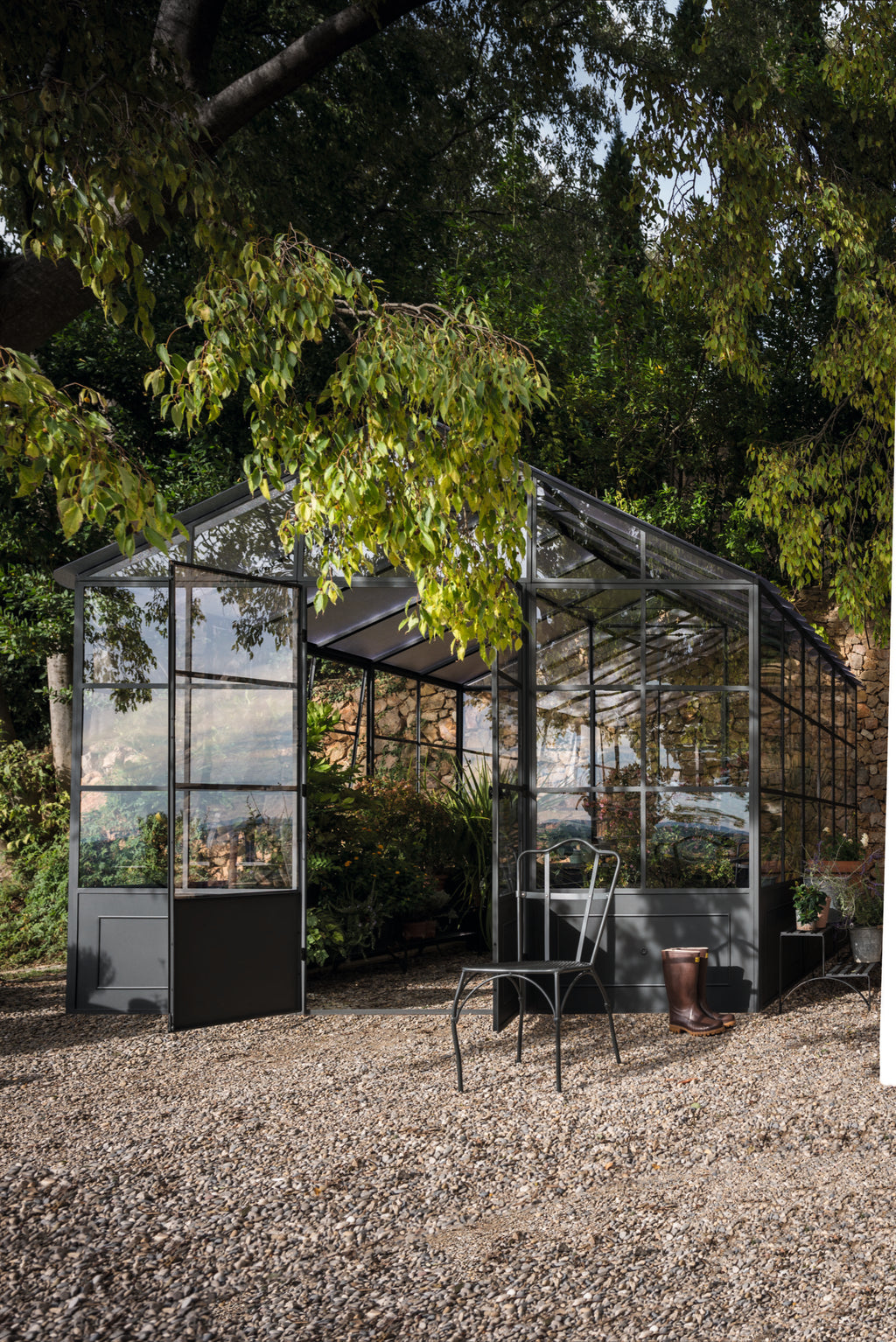 Eine freistehende Orangerie aus Glas und Metall liegt eingebettet zwischen üppigen Bäumen und Laub. Im Inneren der Orangerie gedeihen verschiedene Pflanzen. Draußen stehen ein Metallstuhl und eine kleine Gießkanne auf einer Kiesfläche. Die Szene wird durch natürliches Sonnenlicht beleuchtet.