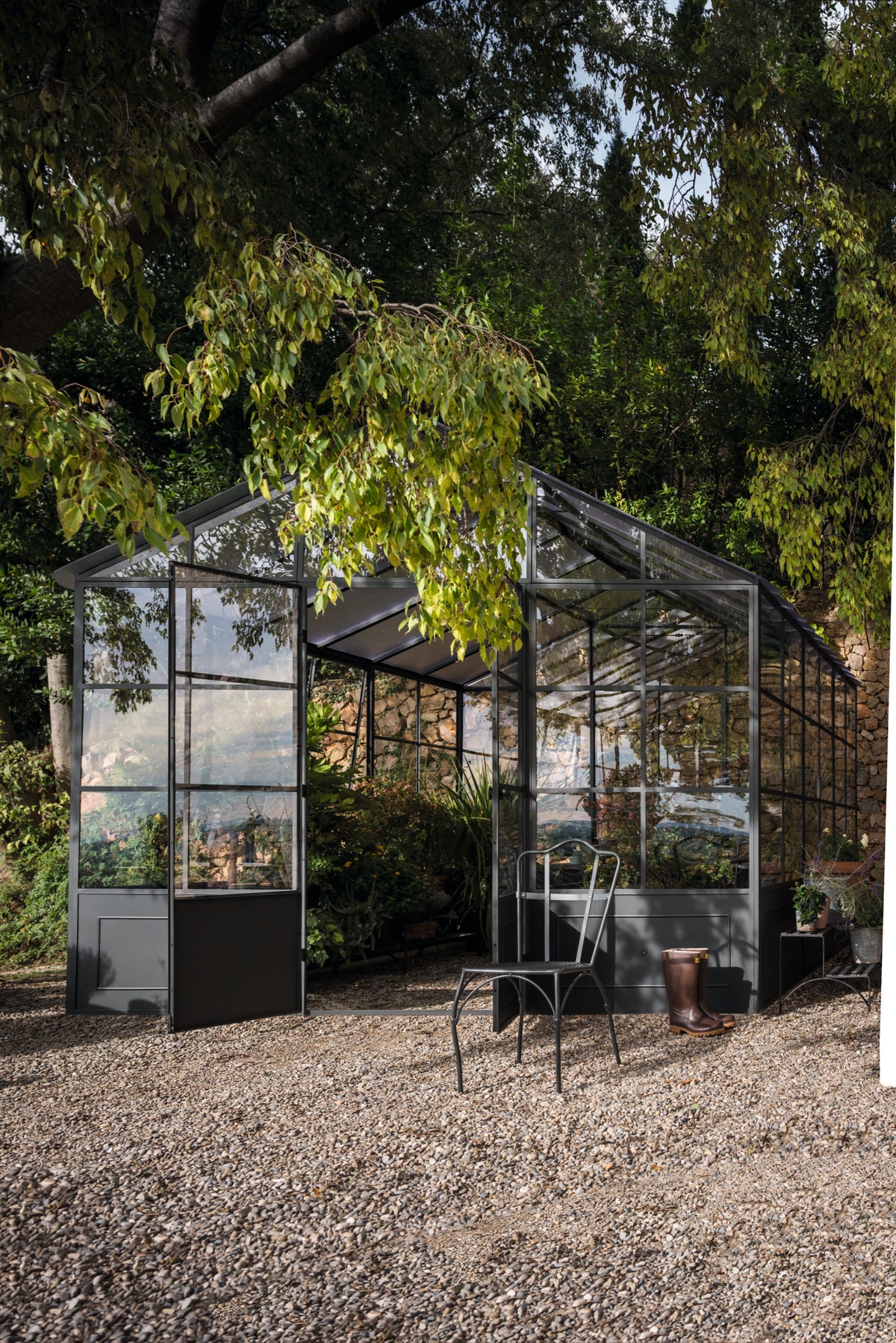 Eine freistehende Orangerie aus Glas und Metall liegt eingebettet zwischen üppigen Bäumen und Laub. Im Inneren der Orangerie gedeihen verschiedene Pflanzen. Draußen stehen ein Metallstuhl und eine kleine Gießkanne auf einer Kiesfläche. Die Szene wird durch natürliches Sonnenlicht beleuchtet.