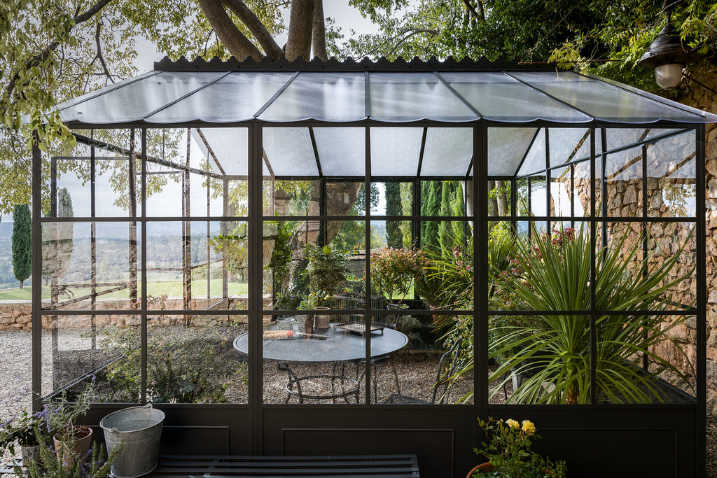 In einem ruhigen Garten steht eine kleine freistehende Orangerie mit großen Fenstern und einem Wellblechdach. Im Inneren sind ein paar Stühle und ein runder Tisch von verschiedenen Pflanzen umgeben. Draußen im Garten führen Steinmauern und Kieswege zu einer entfernten, malerischen Landschaft.