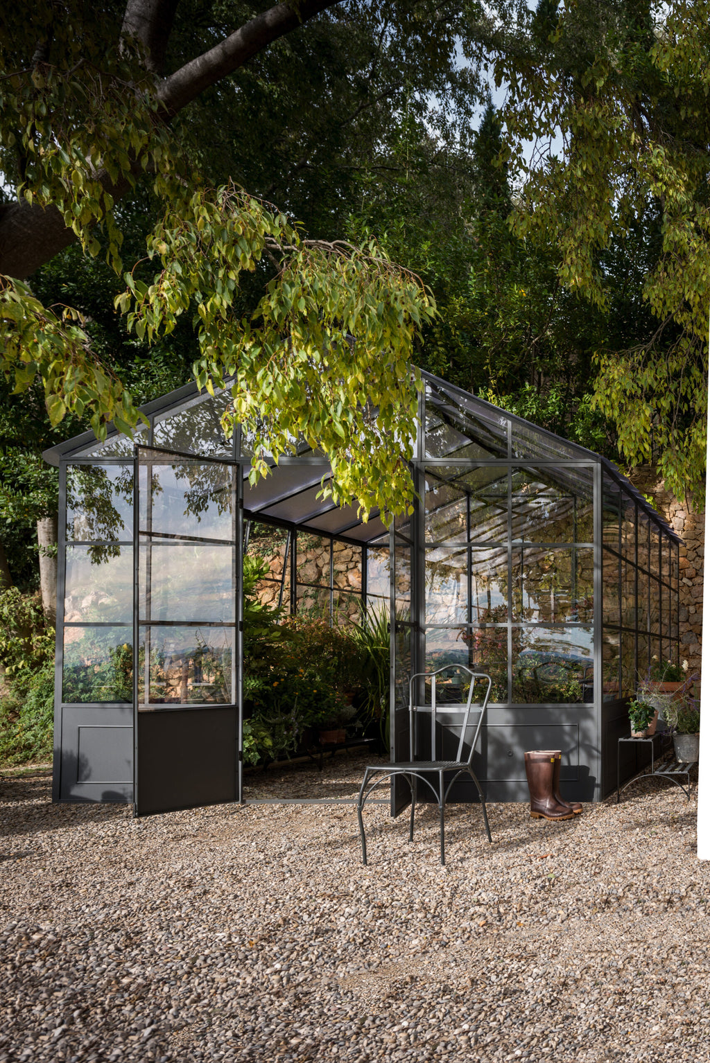 Eine freistehende Orangerie mit großen Glasflächen steht inmitten üppigen Grüns. Die Tür ist geöffnet und gibt den Blick auf verschiedene Pflanzen im Inneren frei. Davor stehen ein Metallstuhl und eine Topfpflanze auf einem Kiesweg. Im Schatten der nahen Bäume wirkt die Umgebung ruhig und einladend und erinnert an eine Orangerie.