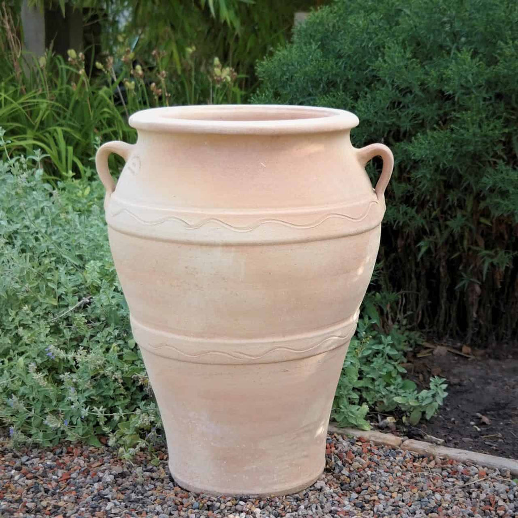 Die frostfeste Terrakotta-Amphore „Olea“ (H=50 cm), eine große beige Urne mit zwei seitlichen Griffen und wellenförmigem Dekormuster um den Korpus, steht auf einer Kiesfläche inmitten grüner Blätter in einer Gartenanlage im Freien.
