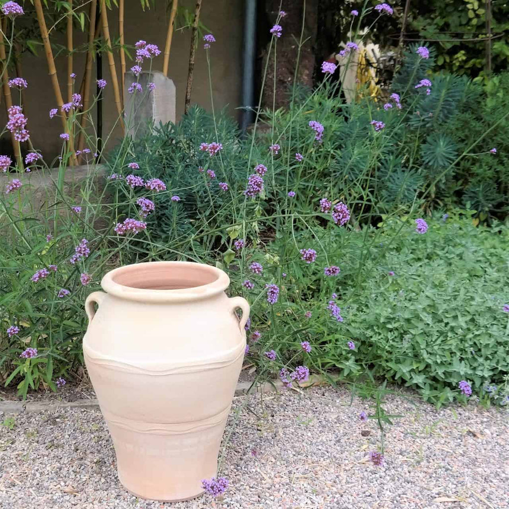 Eine beige Keramikvase mit zwei Griffen steht auf einem Kiesweg vor einem üppigen Gartenbeet voller Grünpflanzen und Büscheln kleiner, zarter lila Blüten. Im Hintergrund sind hohe Bambusstäbe und weiteres Grün zu sehen.