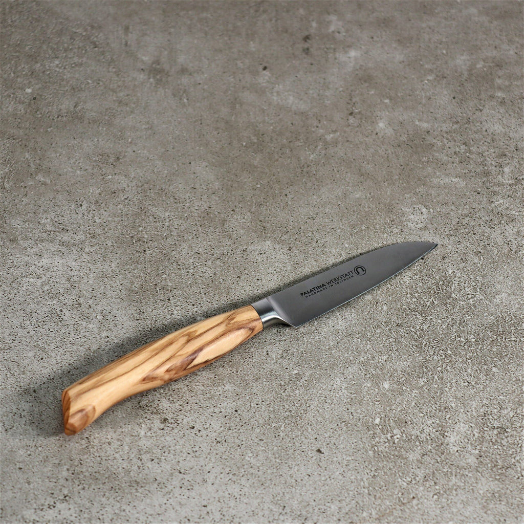 Ein kleines Gemüsemesser der Palatina Werkstatt Messerserie mit Olivenholzgriff und 9 cm langer Klinge aus rostfreiem Stahl mit einzigartiger Maserung ruht auf einer strukturierten Betonoberfläche. Das Markenlogo und der Name sind auf der Klinge sichtbar.