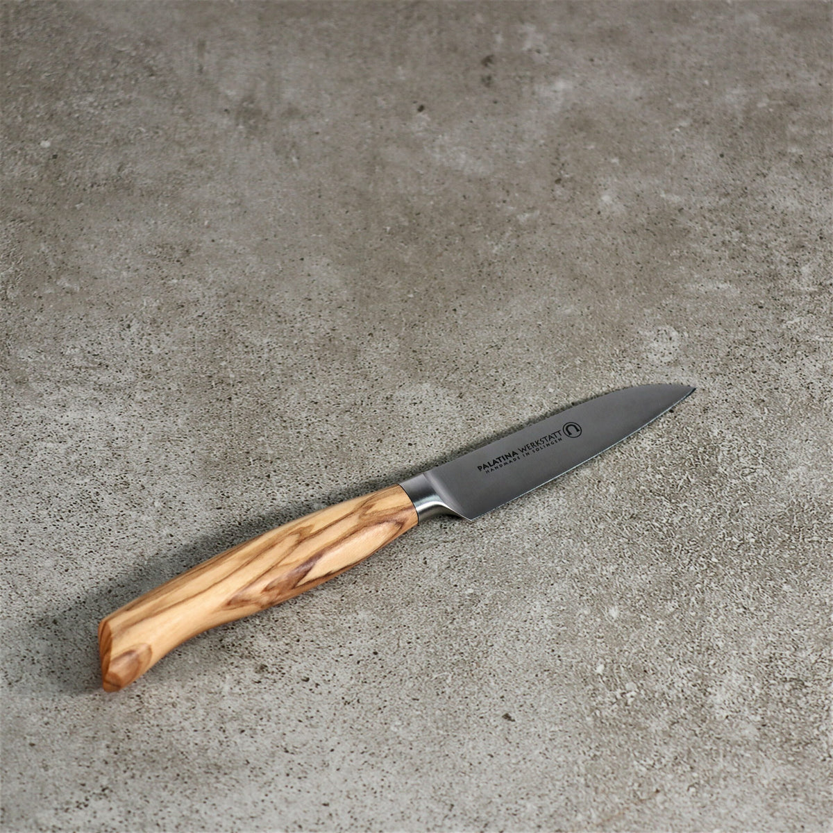 Ein kleines Gemüsemesser der Palatina Werkstatt Messerserie mit Olivenholzgriff und 9 cm langer Klinge aus rostfreiem Stahl mit einzigartiger Maserung ruht auf einer strukturierten Betonoberfläche. Das Markenlogo und der Name sind auf der Klinge sichtbar.