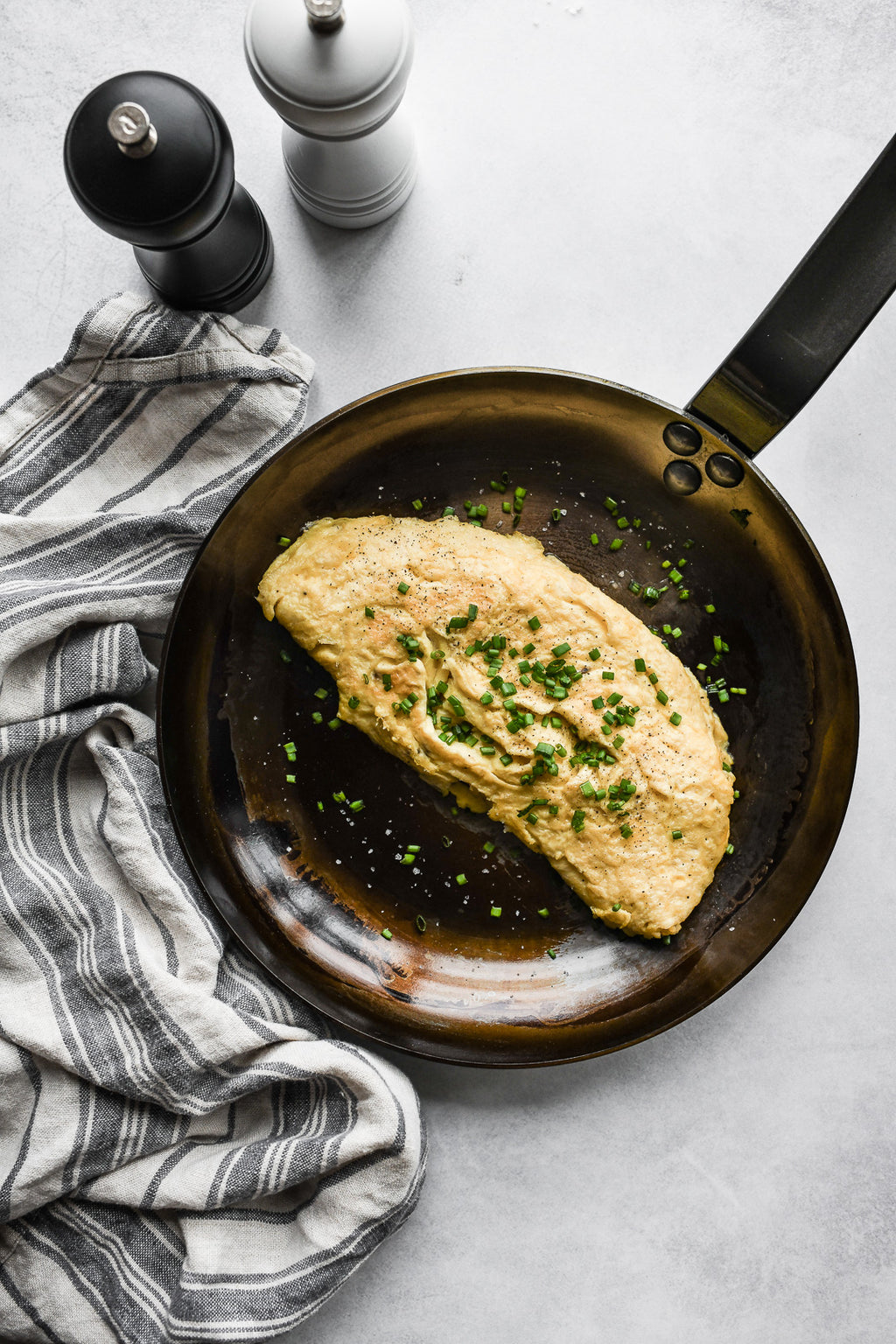 Ein gekochtes Omelett mit Schnittlauch liegt in einer De Buyer Omelette-Pfanne Mineral B, 24 cm Dm, neben einem gestreiften Küchentuch und schwarzen und weißen Pfeffermühlen auf einer hellen Fläche.