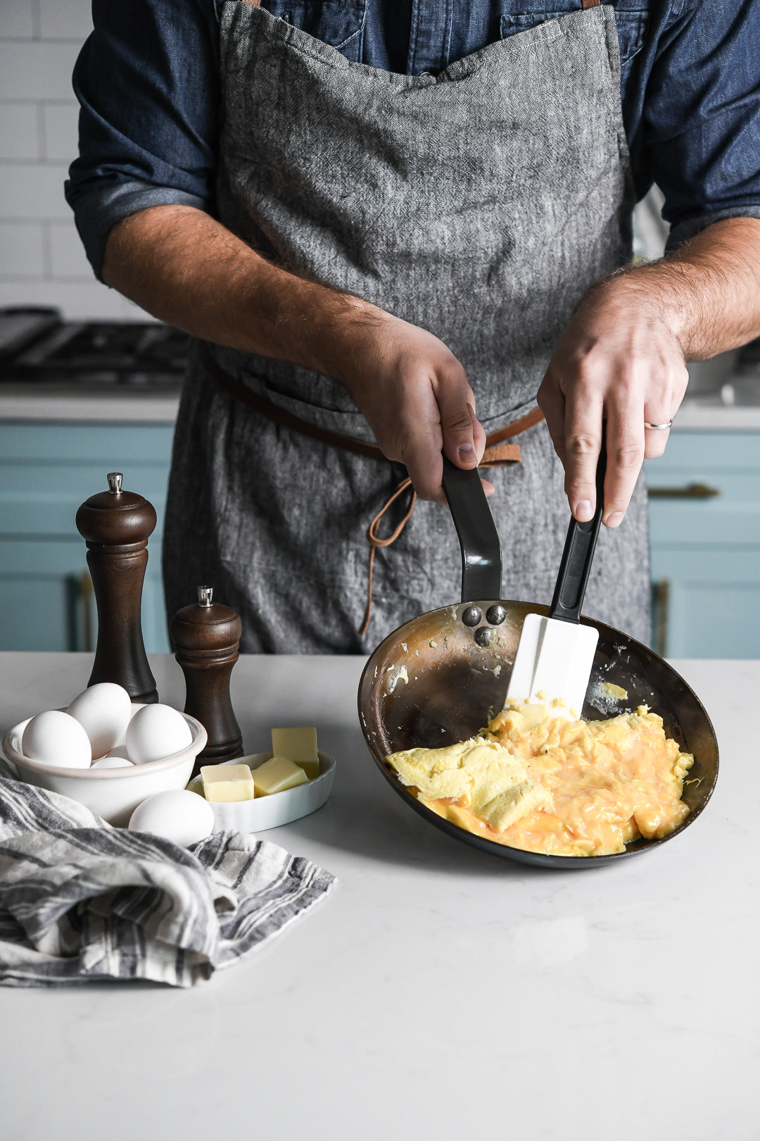 Eine Person in einer grauen Schürze kocht mit einem Spachtel Rührei in einer De Buyer Omelette-Pfanne Mineral B, 24 cm Dm. Eier, Butter, Salz- und Pfefferstreuer und ein gestreiftes Handtuch liegen auf dem Tresen.