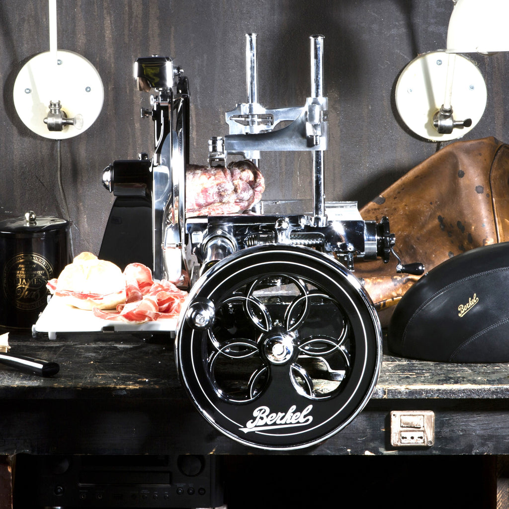 Eine alte Berkel Volano P15 Schwarz Aufschnittmaschine mit Schwungrad+ Standfuß und Aufschnittbrett steht auf einer dunklen Holztheke mit aufwendigen schwarzen und verchromten Details. Aufgeschnittenes Fleisch wird auf einem weißen Tablett daneben präsentiert. Im Hintergrund sind zwei runde Lampen und verschiedene Lederartikel zu sehen. Auf der Aufschnittmaschine ist das Berkel-Logo zu sehen.