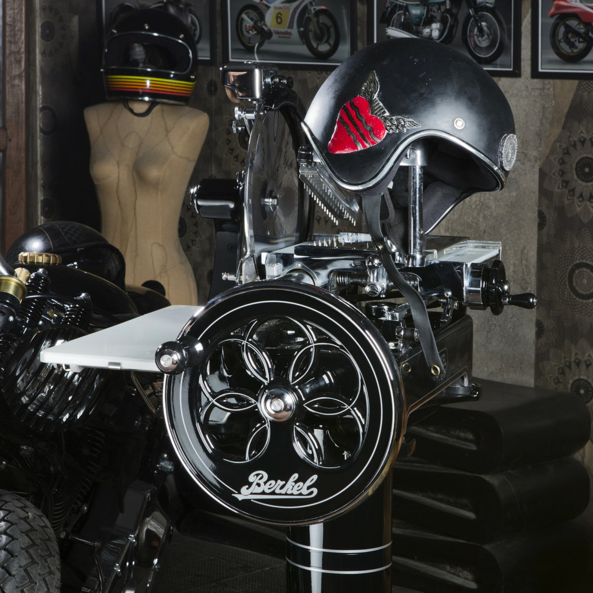 Eine alte schwarze Berkel Volano P15 Aufschnittmaschine mit Schwungrad+ Standfuß und Aufschnittbrett steht prominent in einem Raum, der mit verschiedenen Motorradhelmen geschmückt ist. Ein schwarzer Helm mit rotem Design thront auf der Aufschnittmaschine, während im Hintergrund eine Schaufensterpuppenbüste zu sehen ist, die zur eklektischen Atmosphäre beiträgt.