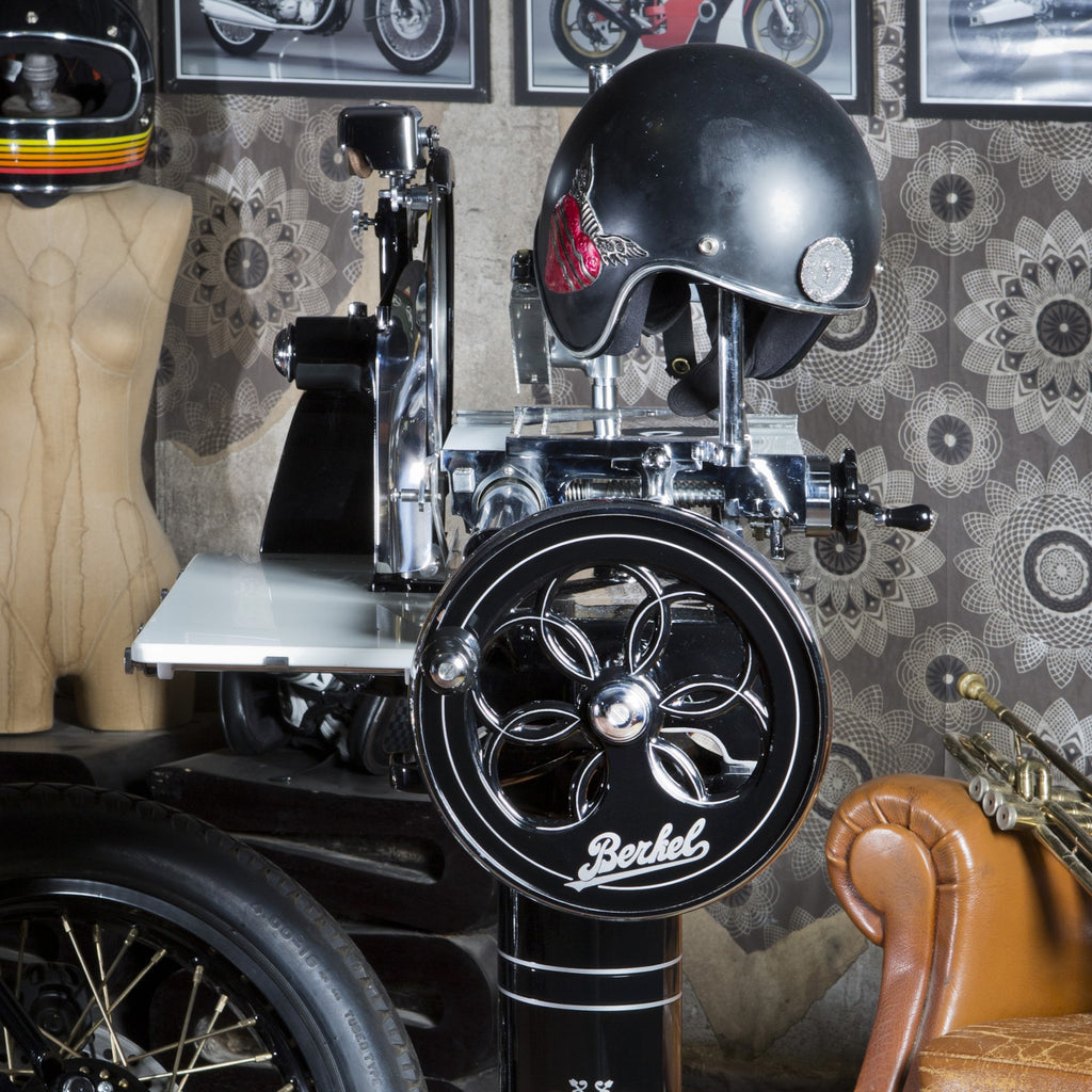 Eine schwarze Berkel Volano P15 Aufschnittmaschine mit Schwungrad + Standfuß und Aufschnittbrett, geschmückt mit einem schwarzen Motorradhelm, steht in einem Raum voller verschiedener Gegenstände wie einer Schaufensterpuppe und dekorativer Wandkunst. Auf der linken Seite des Bildes ist teilweise ein Motorradrad sichtbar, während auf der rechten Seite ein brauner Lederstuhl steht.