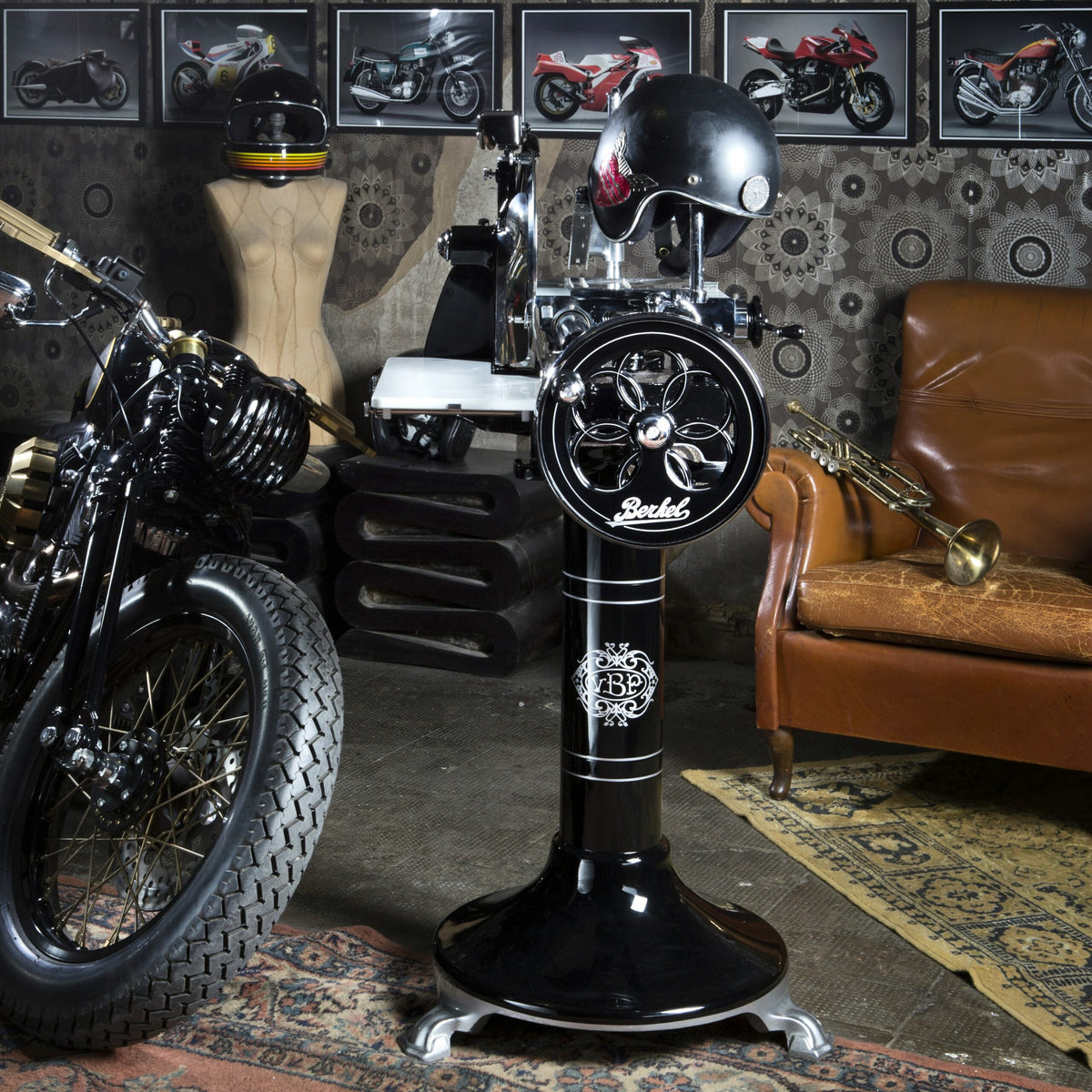 Ein Raum mit Vintage-Motorradthema verfügt über einen schwarzen Motorradsitz aus Leder, einen klassischen schwarzen Friseurstuhl mit einem Helm darauf, gerahmte Motorradfotos an der Wand, einen braunen Ledersessel und im Hintergrund eine schwarze Berkel Volano P15 Aufschnittmaschine mit Schwungrad + Standfuß und Aufschnittbrett. Der Boden ist mit gemusterten Teppichen bedeckt.