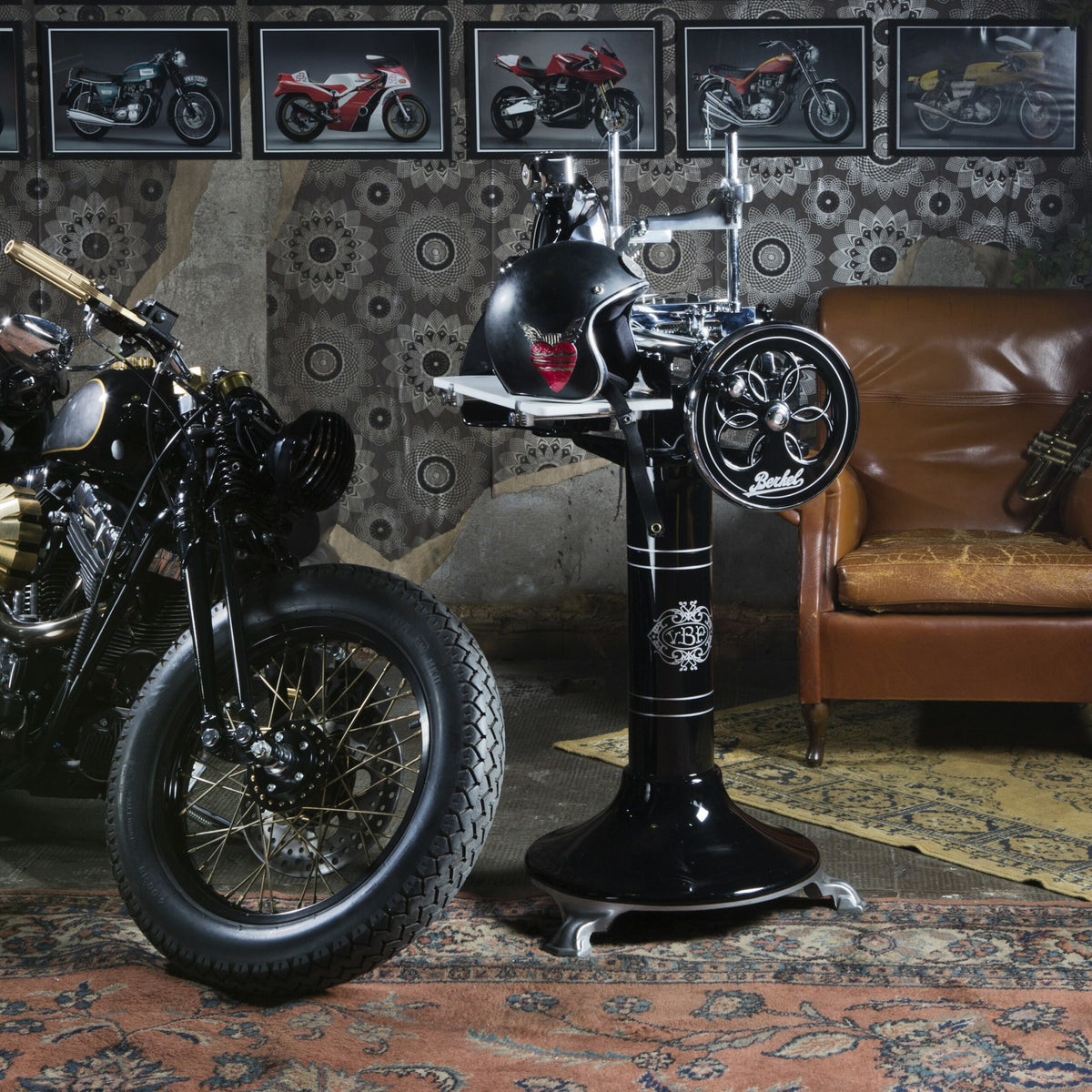 In einem Raum im Vintage-Stil stehen Motorräder, darunter eines mit einer Berkel Volano P15 schwarz Aufschnittmaschine mit Schwungrad+ Standfuß und Aufschnittbrett und aufwendigen Motordesigns, neben einem stilvollen schwarzen Friseurstuhl mit Helmhalter und Retro-Friseurwerkzeugen. Gerahmte Fotos von Motorrädern zieren die gemusterte Tapete und tragen zum Old-School-Ambiente bei.
