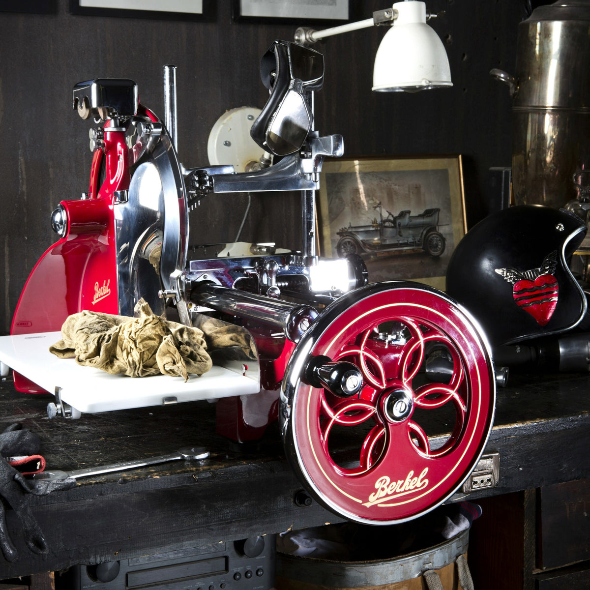 Eine alte Berkel Volano P15 Rot Aufschnittmaschine Schwungrad, komplett mit Ständer und Schneidebrett, steht auf einer dunklen Holztheke. Über die Arbeitsfläche der Aufschnittmaschine ist ein Tuch drapiert. Im Hintergrund sind ein monochromes Foto eines Oldtimers, ein schwarzer Helm mit Herzdesign und verschiedene metallische Objekte zu sehen.