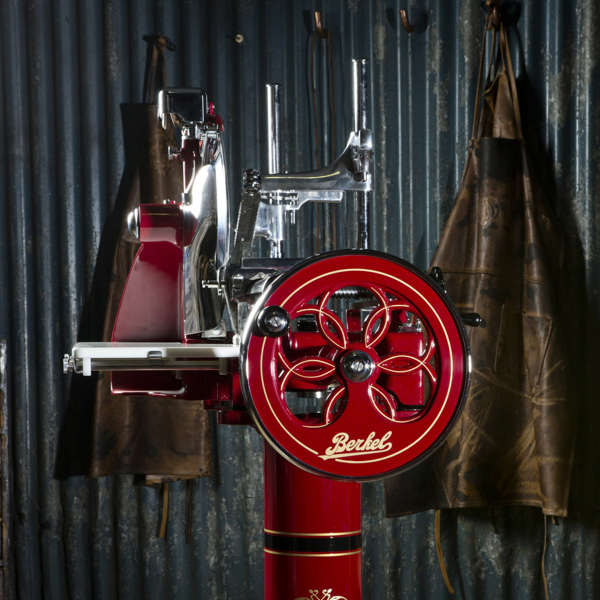 Ein Vintage-Fleischschneider Berkel Volano P15 Rot mit aufwendigen Metalldetails und einem großen, prominent präsentierten Schwungrad. Vor einem Hintergrund aus Wellblech mit zwei an der Wand hängenden braunen Lederschürzen verfügt diese exquisite Aufschnittmaschine über einen eleganten Standfuß für Stabilität und ein Aufschnittbrett für Komfort.