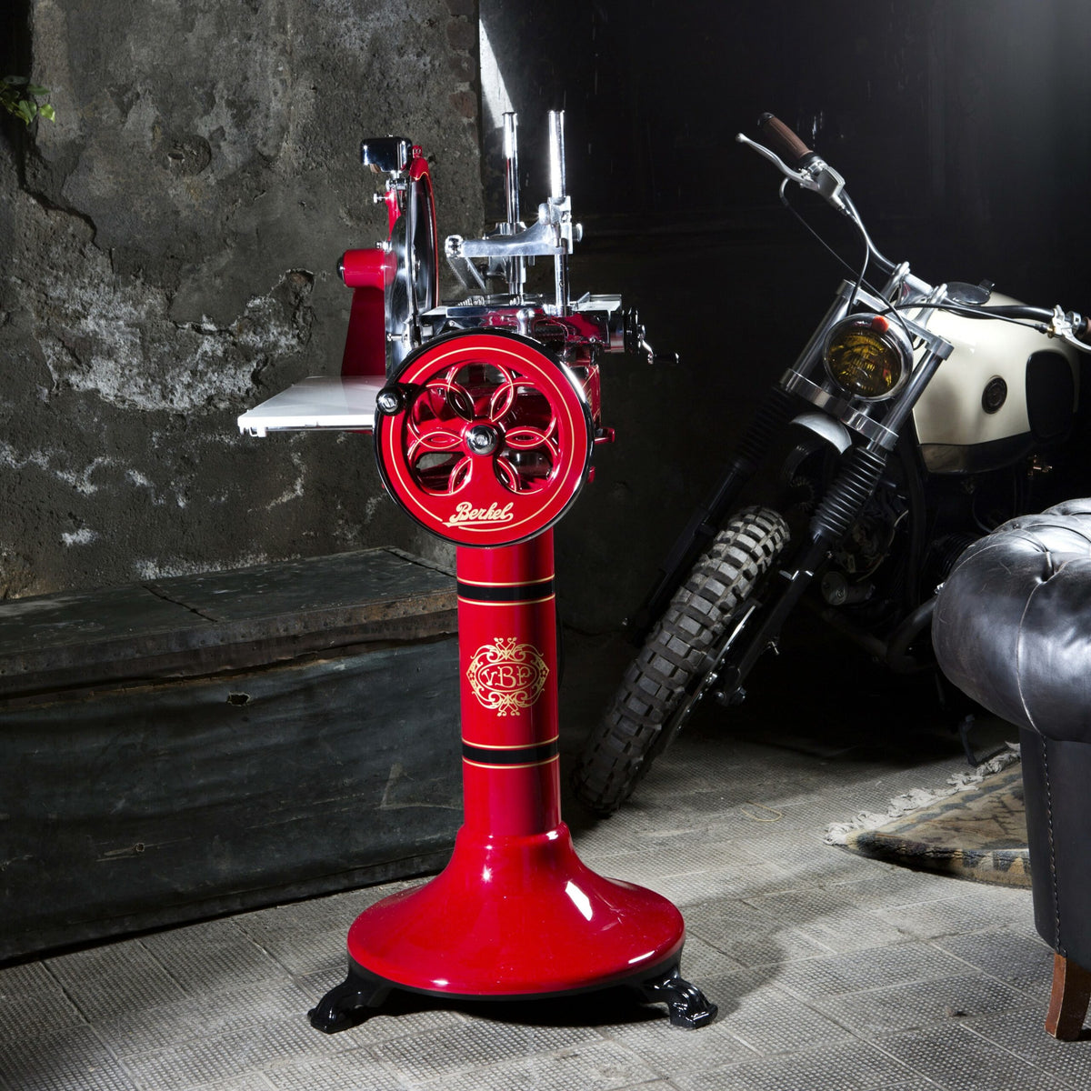 Ein Vintage-Fleischschneider Berkel Volano P15 Rot mit kunstvollen Details steht auf einem gemusterten Boden vor einer Betonwand mit verwitterter Optik. Dahinter steht ein klassisches Motorrad mit rundem Scheinwerfer und rechts ein abgedunkeltes Ledersofa. Die Umgebung strahlt eine industrielle Atmosphäre aus.