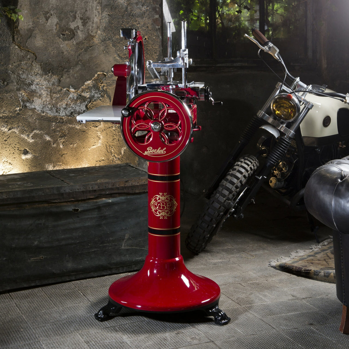 Eine Vintage-Aufschnittmaschine für rotes Fleisch, die edle Berkel Volano P15 Rot Aufschnittmaschine Schwungrad mit Standfuß und Aufschnittbrett, steht prominent in einem rustikalen Raum mit strukturierten Betonwänden und gedämpfter Beleuchtung. Dahinter parkt ein schwarz-weißes Motorrad. Die Umgebung strahlt einen industriellen und Retro-Charme aus und zeigt die zeitlose Eleganz des Schwungrad-Designs.