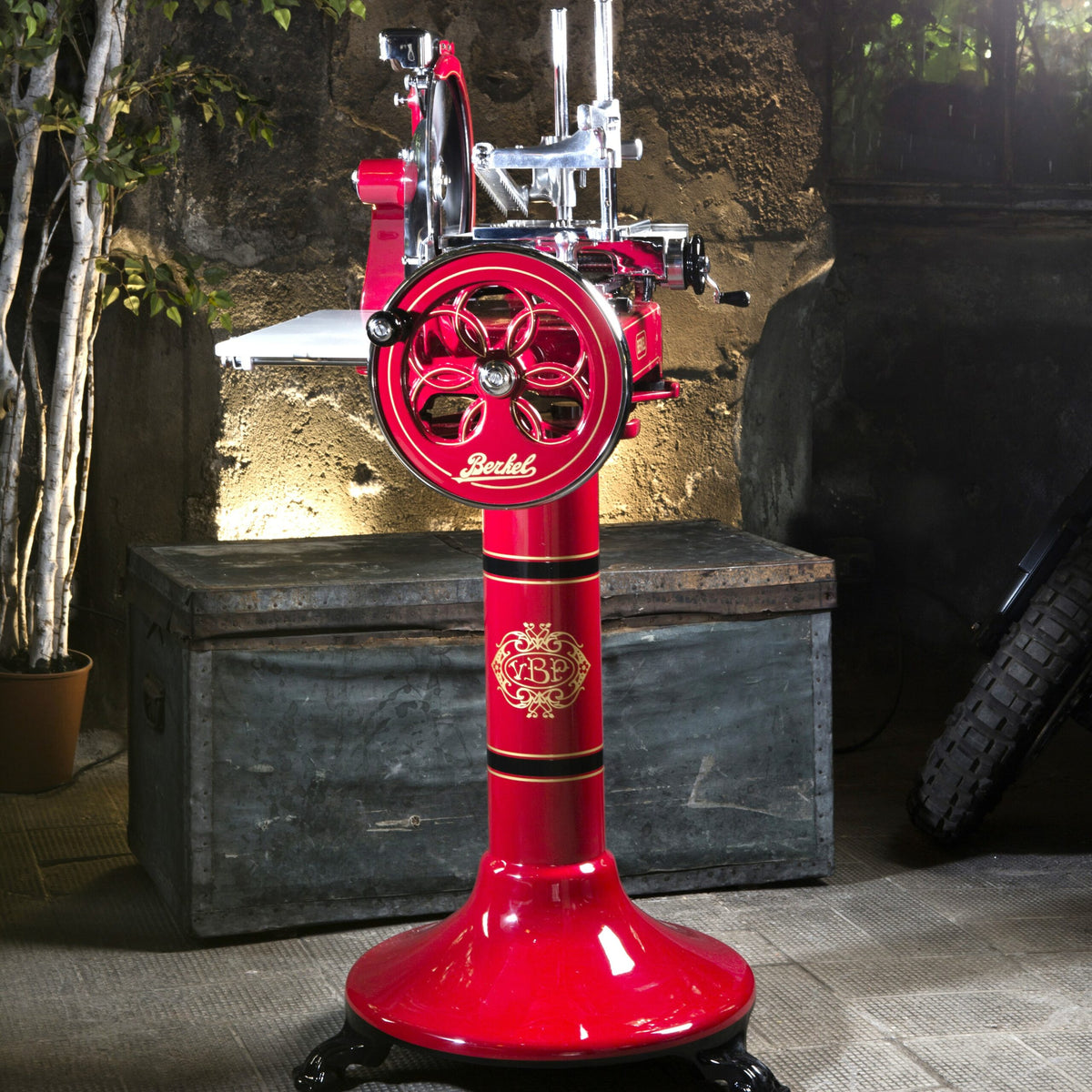 Eine Berkel Volano P15 Rot, Aufschnittmaschine Schwungrad + Standfuß + Aufschnittbrett, im antiken Stil mit verzierten goldenen Details auf einem Sockel, steht drinnen auf einem Steinboden. Im Hintergrund sind eine rustikale Wand, eine Metalltruhe und einige Blattpflanzen zu sehen. Die lebhafte Farbe und das aufwendige Design des Aufschnittgeräts machen es zu einem Blickfang in der Umgebung.