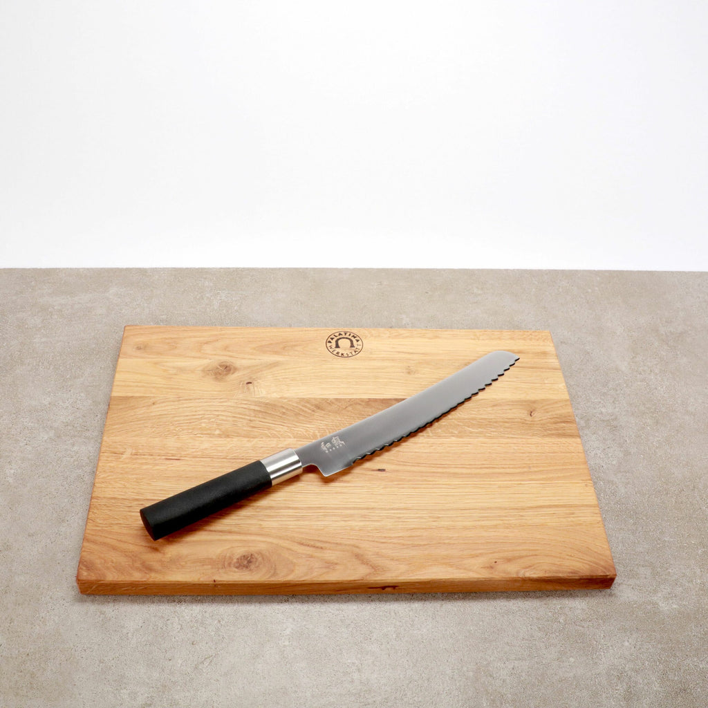 Auf der Oberfläche liegt ein Kai Wasabi Black Brotmesser 23 cm Klinge + großes Eiche Schneidebrett 40x30 cm. Das Schneidebrett hat oben in der Mitte ein kreisförmiges Logo eingraviert. Der Hintergrund ist eine neutrale graue Oberfläche und eine schlichte weiße Wand.