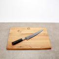 Auf der Oberfläche liegt ein Kai Wasabi Black Brotmesser 23 cm Klinge + großes Eiche Schneidebrett 40x30 cm. Das Schneidebrett hat oben in der Mitte ein kreisförmiges Logo eingraviert. Der Hintergrund ist eine neutrale graue Oberfläche und eine schlichte weiße Wand.