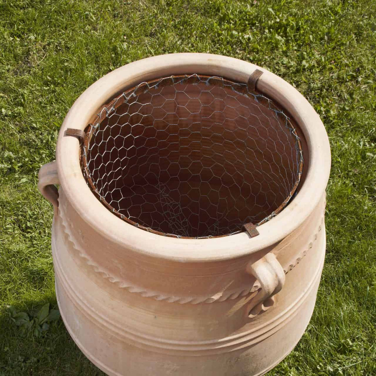 Ein großer Terrakottatopf mit zwei Griffen an den Seiten steht auf Gras und ist oben mit einem Drahtgeflecht bedeckt. Die Oberfläche des Topfes ist mit strukturierten horizontalen Bändern als dekoratives Detail verziert.